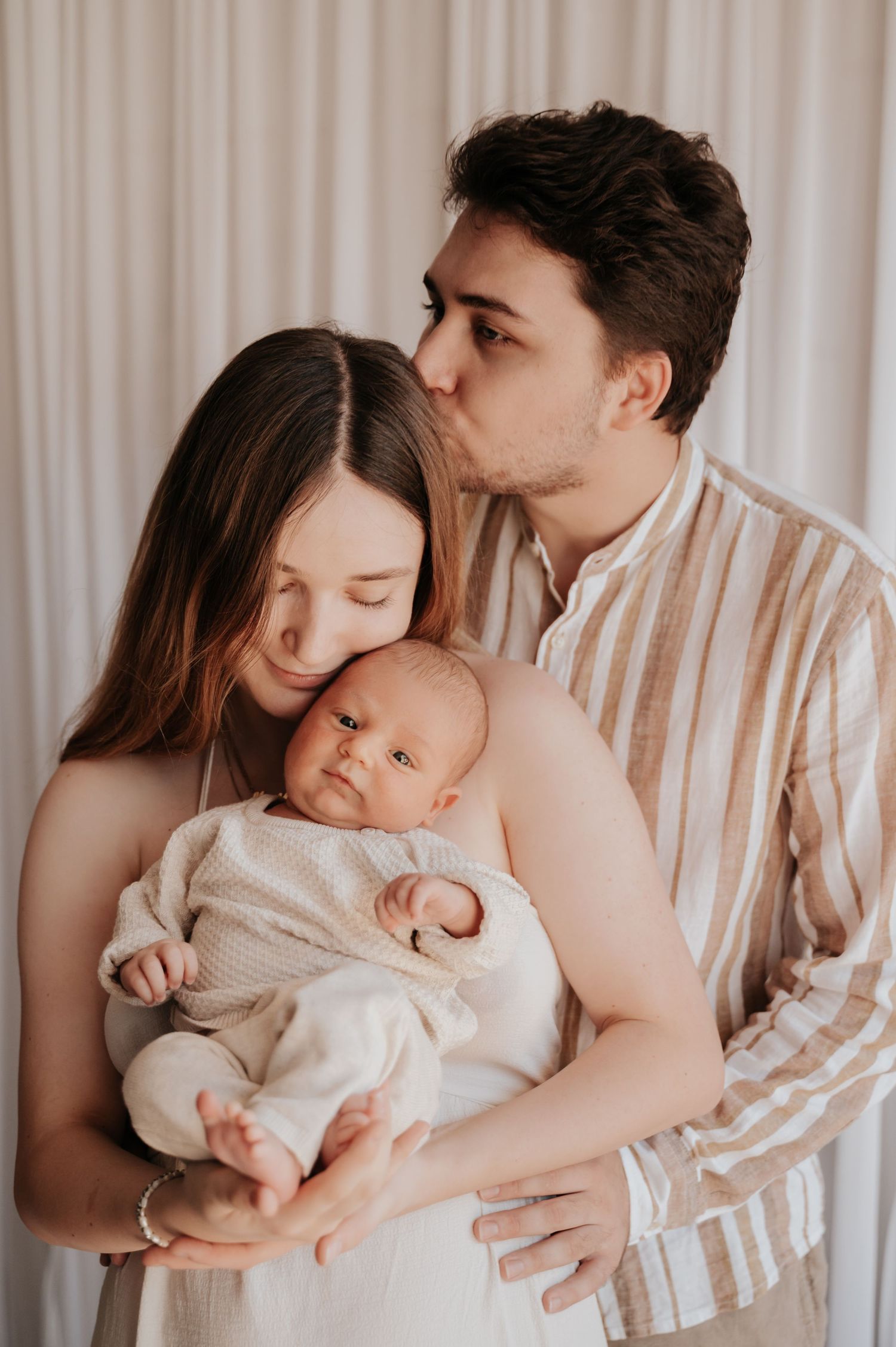Young couple tenderly holding their newborn baby, man kissing woman on head, in soft neutral tones.