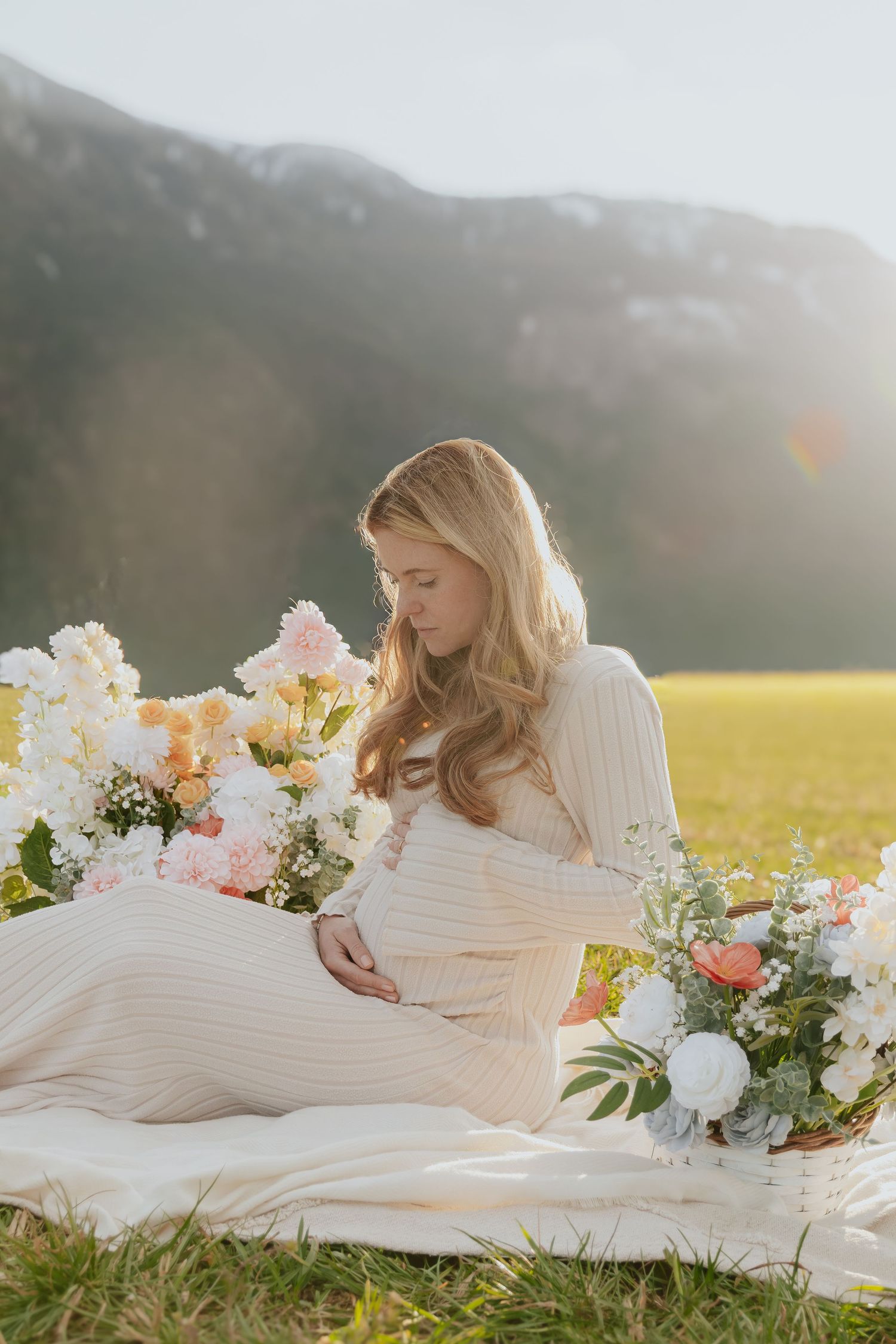 Pregnant woman in cream ribbed dress sitting among flowers with golden mountain backdrop.