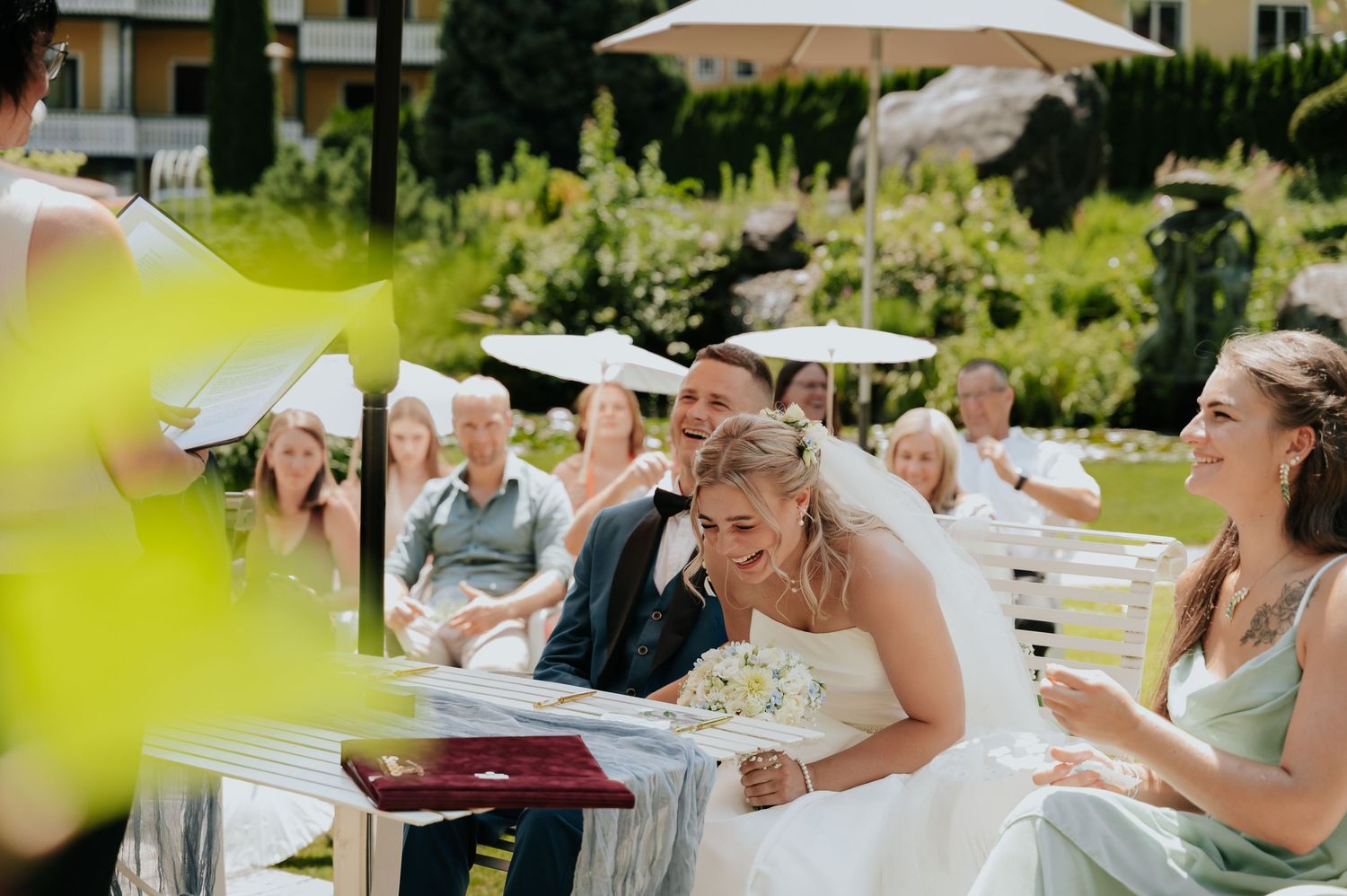 Bride and groom smile during outdoor wedding ceremony with guests seated on white chairs in a sunny garden.