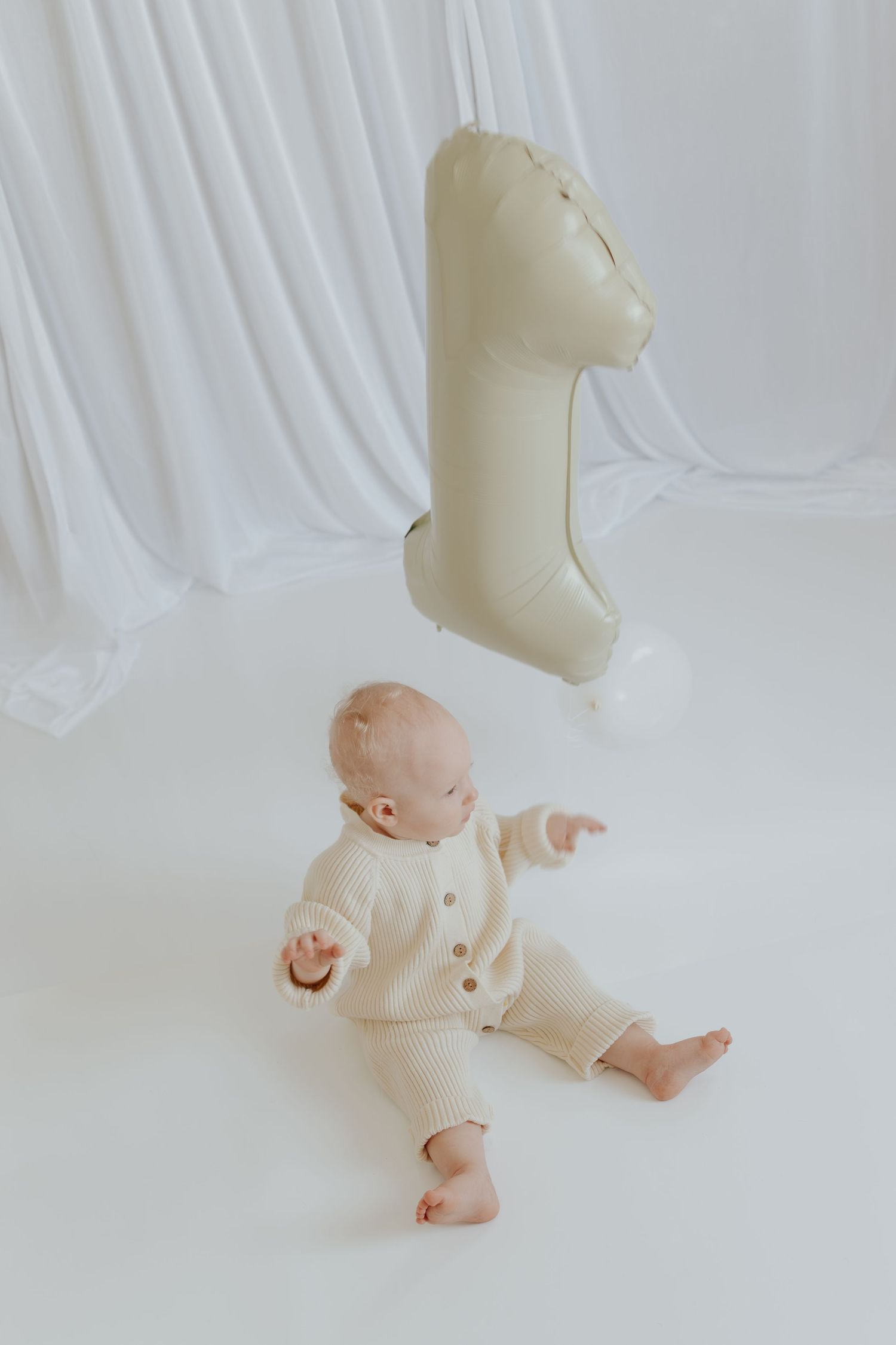 Baby in cream knit outfit sits on white floor reaching toward a number one balloon for first birthday.