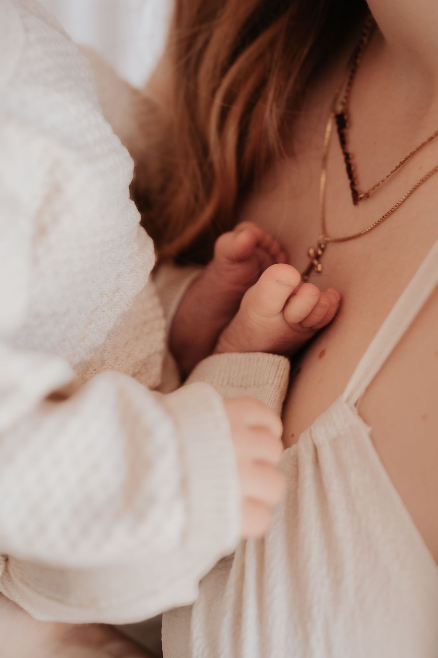 Close-up of a newborn baby's tiny hand grasping a mother's finger, wearing white clothing with gold necklaces visible.