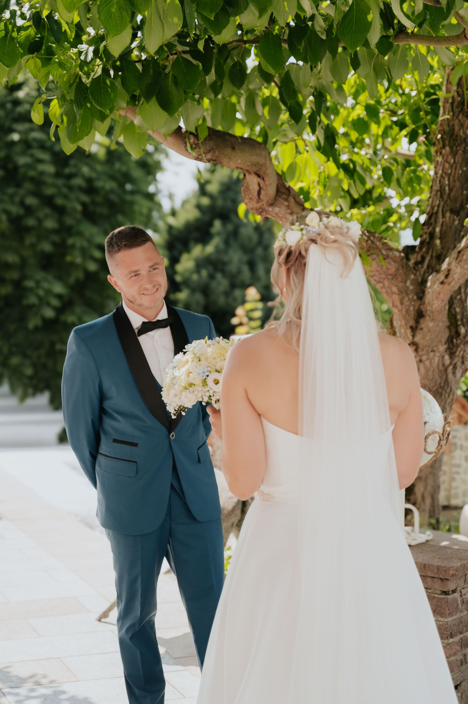 Groom in blue tuxedo smiles at bride in white wedding dress and veil beneath a green tree outdoors.