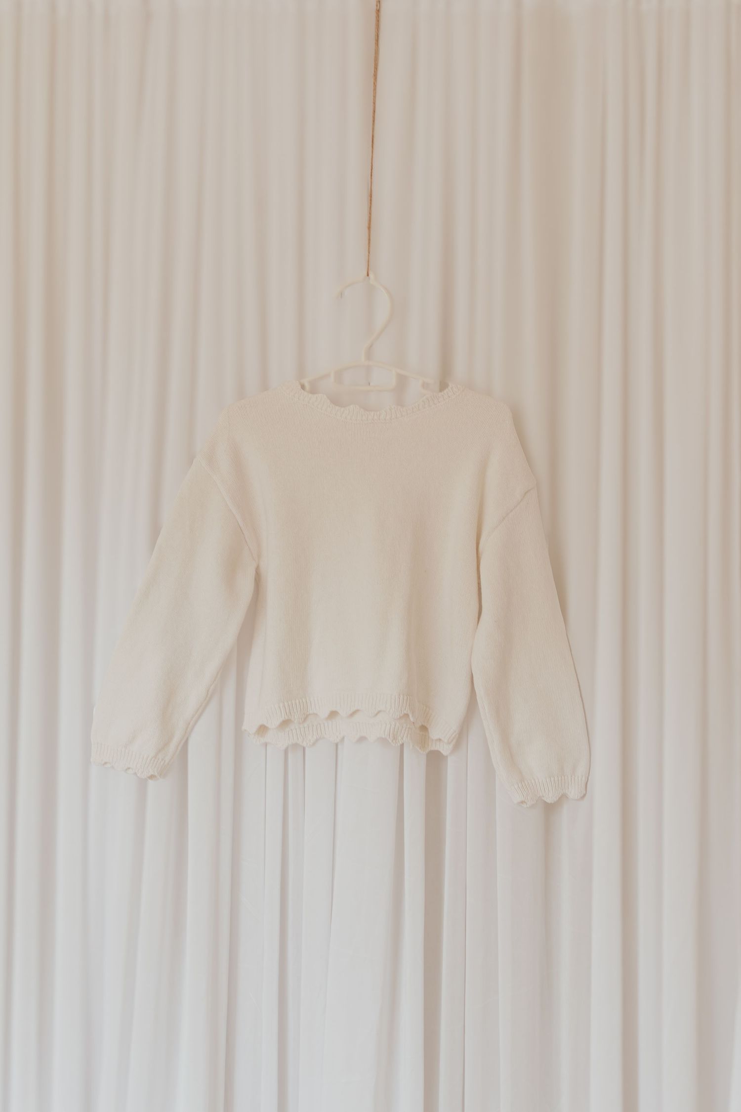 Ein cremefarbenes Sweatshirt, das an einem Kleiderbügel vor einem weißen Vorhanghintergrund hängt.
