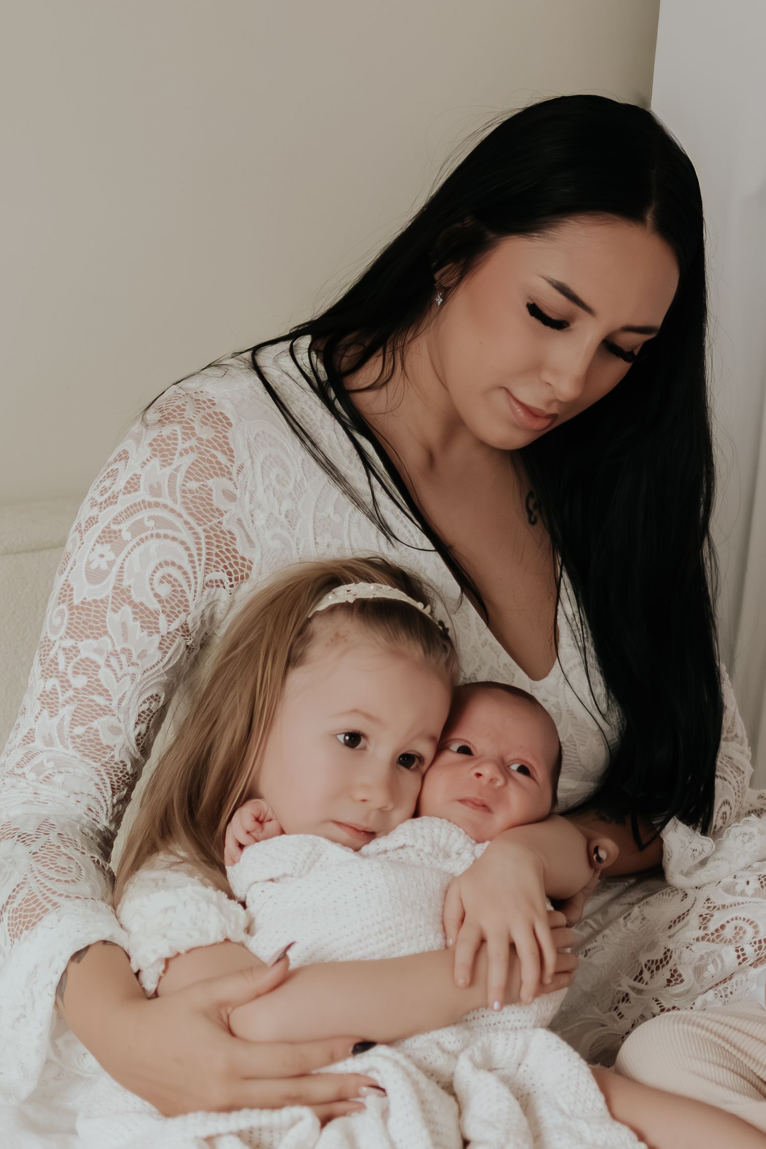 Tender family moment captured in soft white lace as mother embraces two young children in intimate portrait.