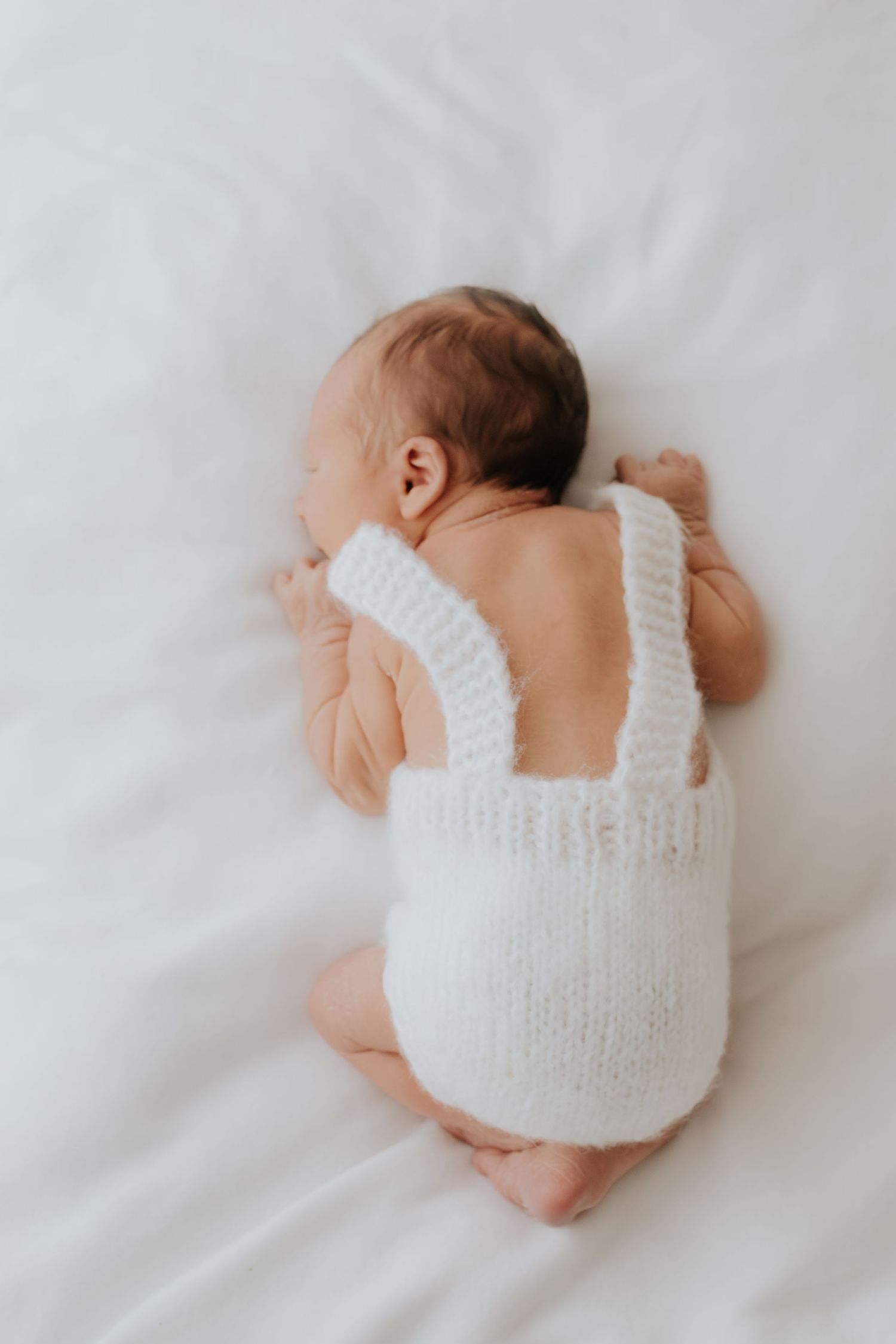 A sleeping newborn baby wearing a white knit diaper cover lies peacefully on white bedding.