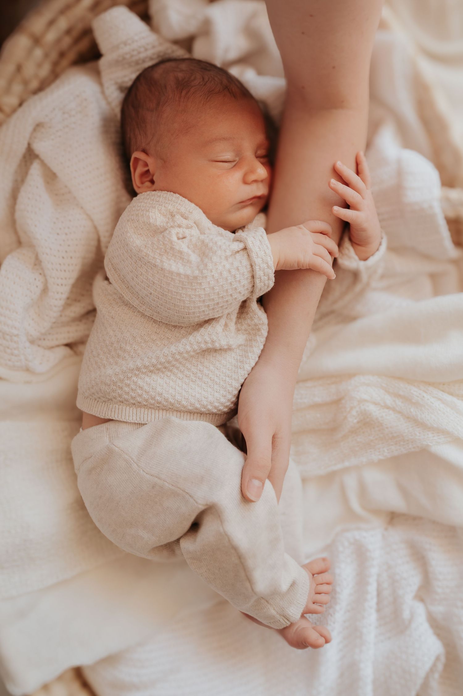 Newborn baby sleeping peacefully in cream knit outfit on white blanket, cradled by mother's arm.