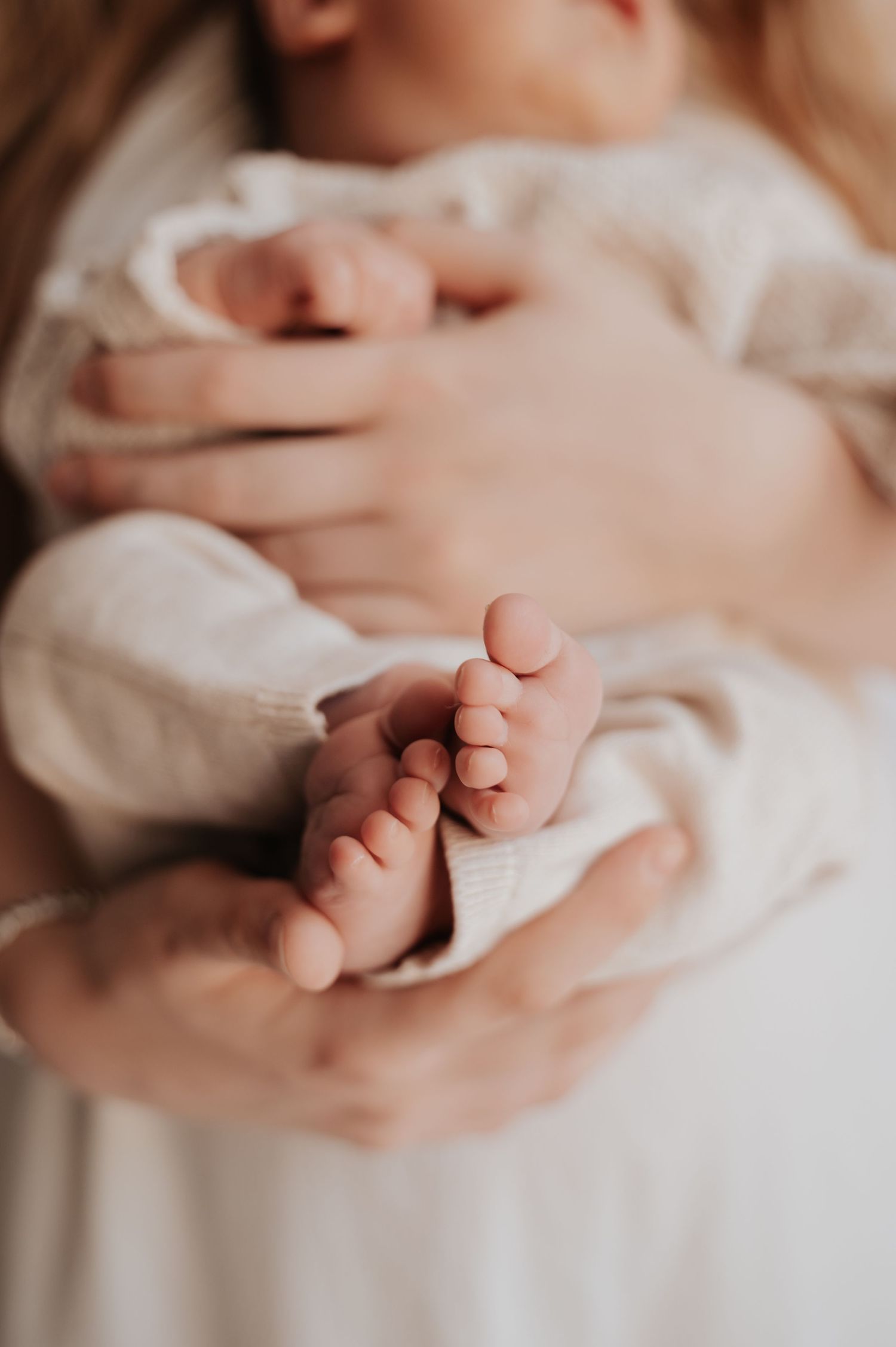 Close-up of adult hands gently cradling tiny newborn baby feet wrapped in a soft white knit blanket.
