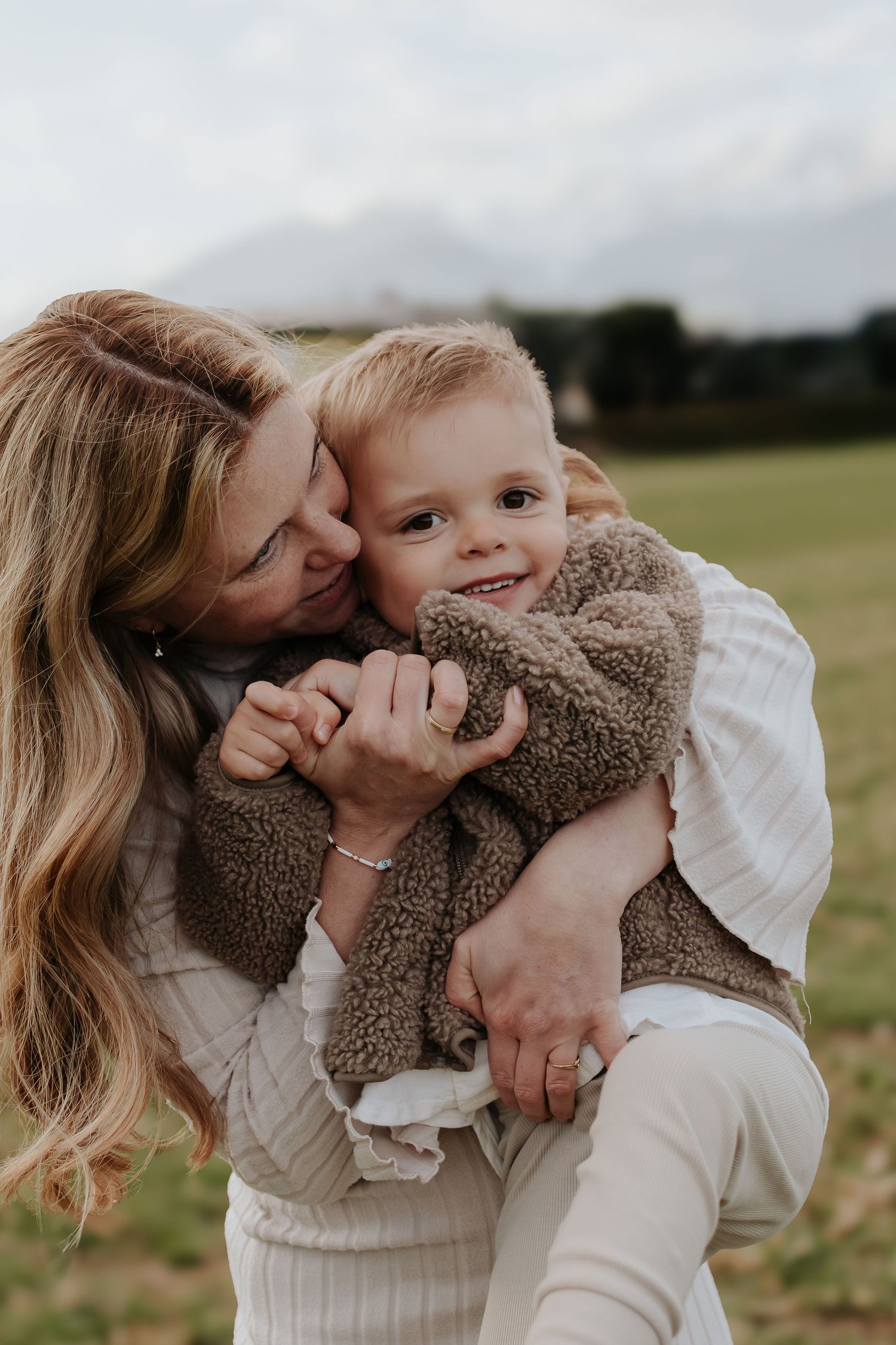 A woman hugging and kissing a smiling toddler boy in a teddy fleece jacket outdoors in a green field.