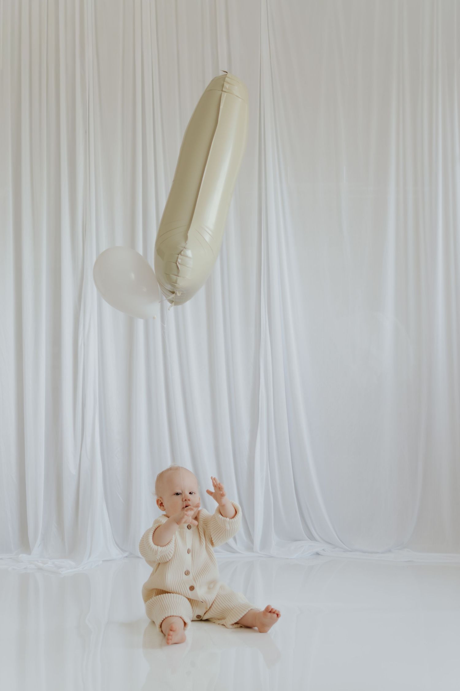 Baby in cream outfit sits on white floor reaching up toward floating number one balloon against white drape backdrop.