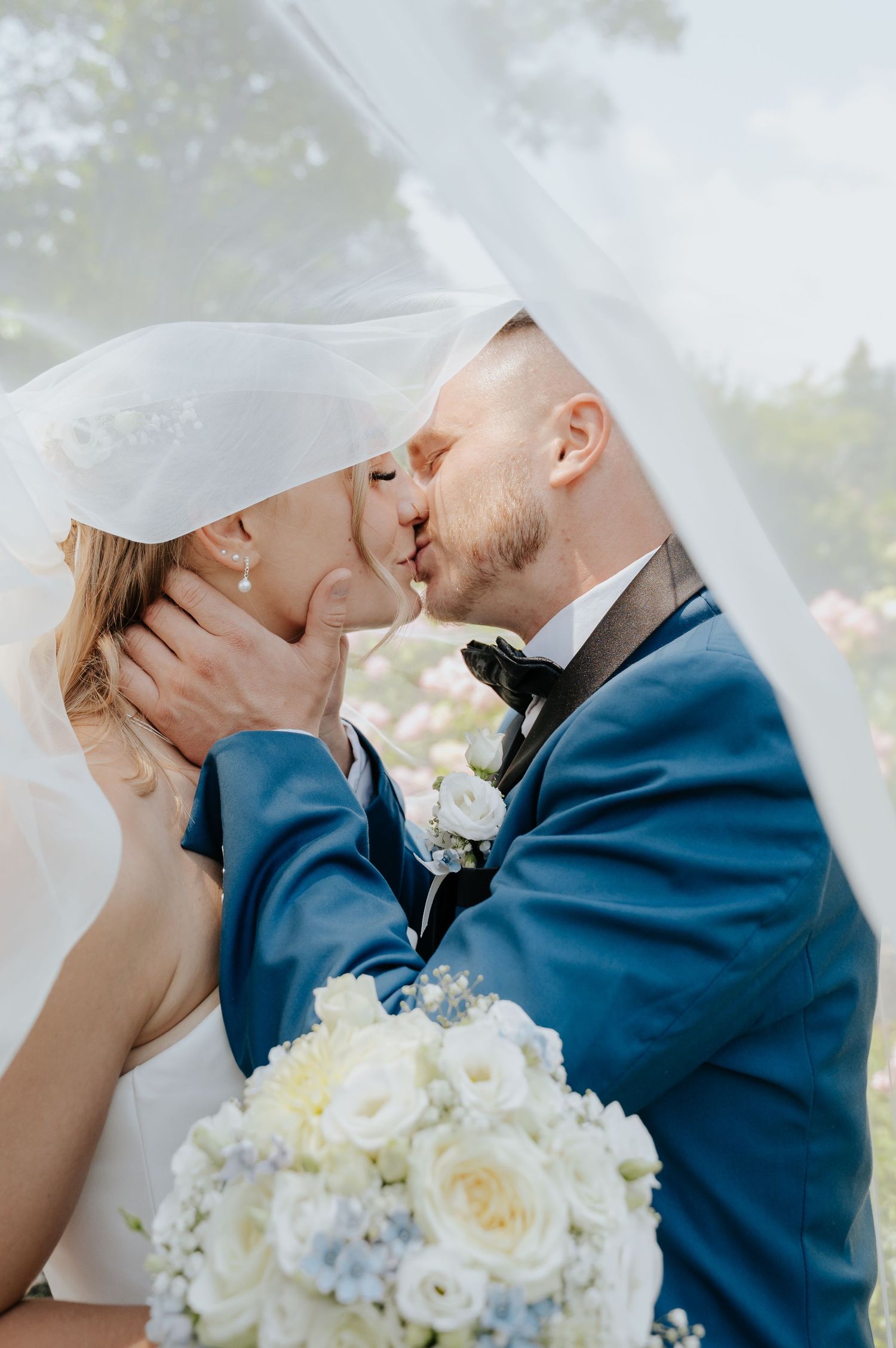 Bride and groom share a romantic kiss under a white veil, with a bouquet of white and blue flowers in the foreground.