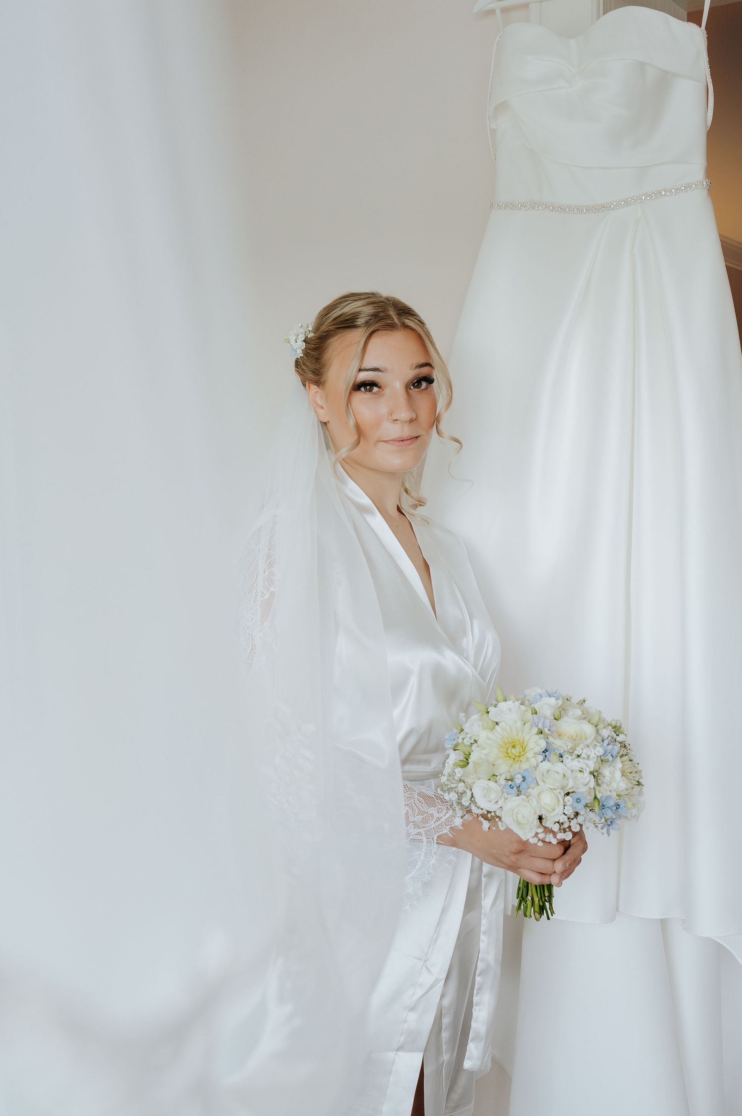 Bride in white satin robe holding elegant bouquet of white and blue flowers, standing beside her wedding gown.