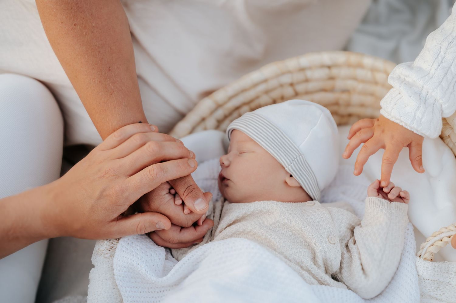 Newborn baby in white outfit and hat resting peacefully while parent gently holds tiny hands in a cozy basket.