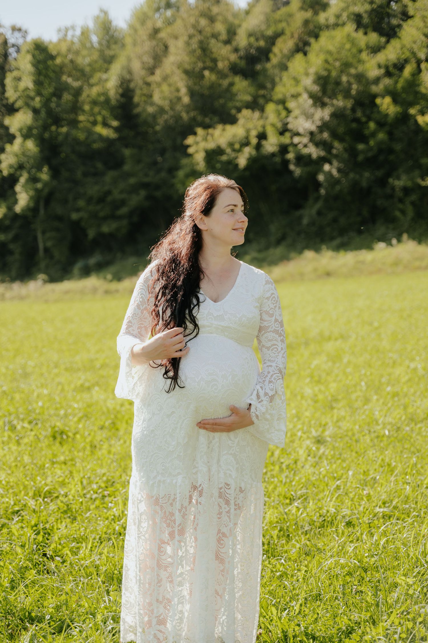 Pregnant woman in white lace dress cradling baby bump in sunny green meadow with trees.