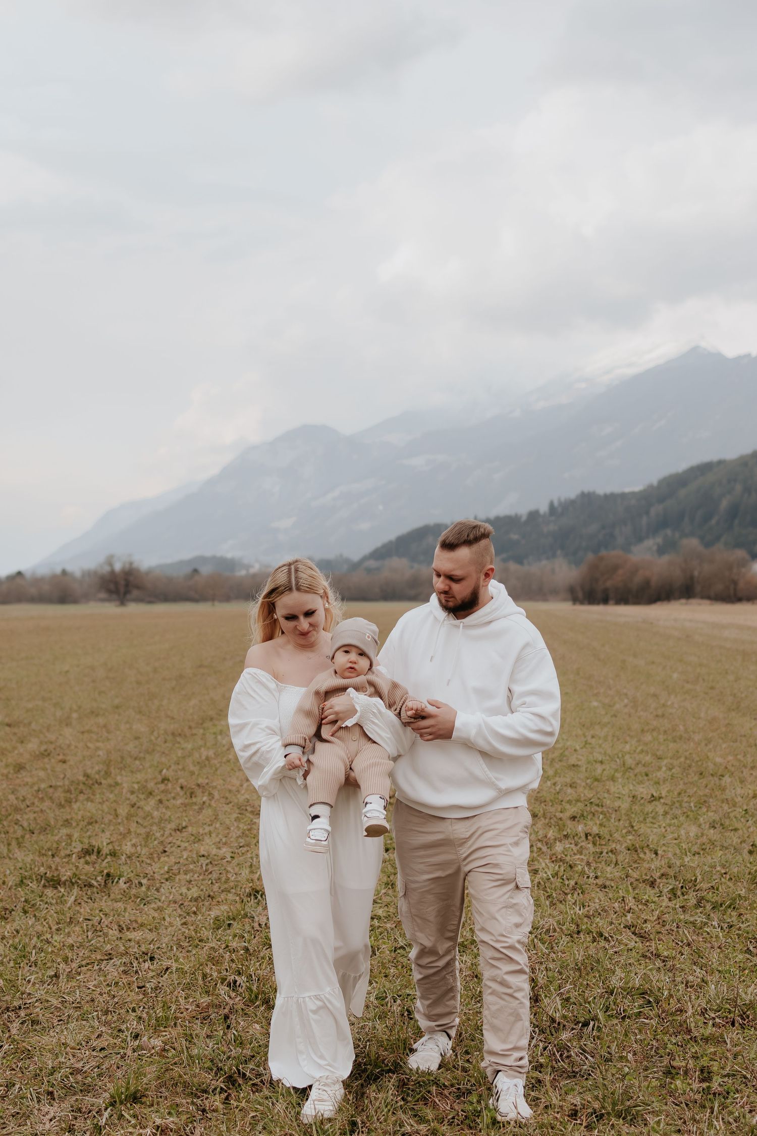 Young family with baby posing in open field with mountain backdrop on cloudy day.