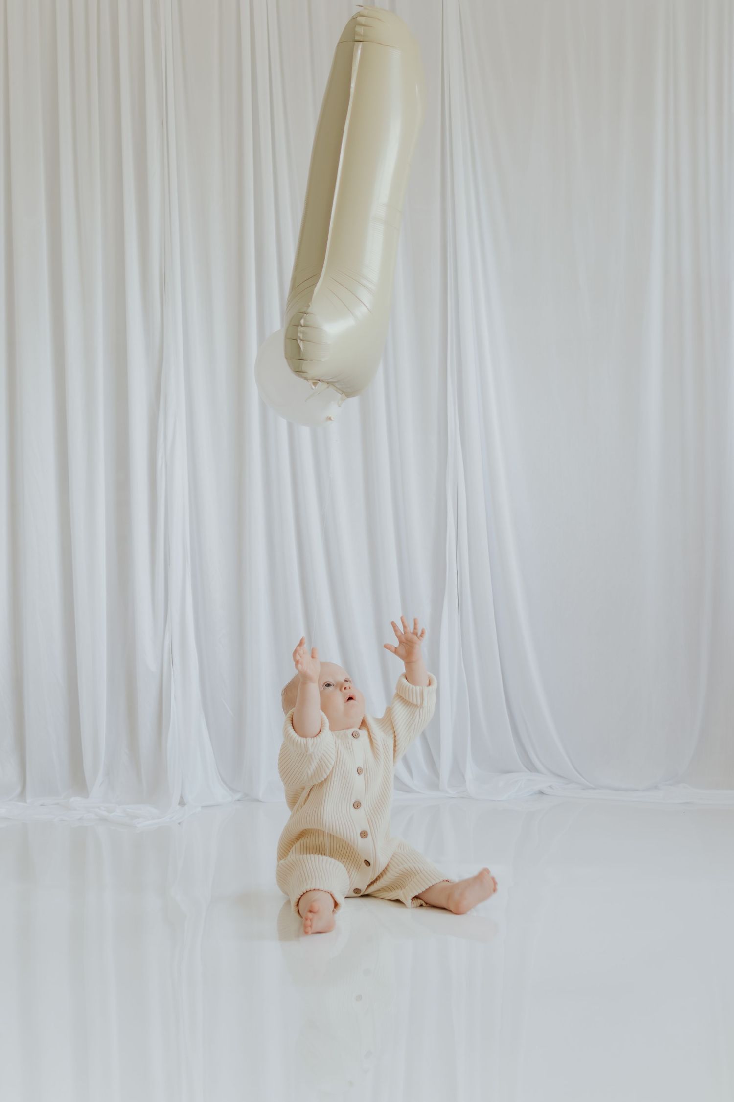 Baby in cream outfit reaches for a number one balloon while sitting on white floor against white draped backdrop.