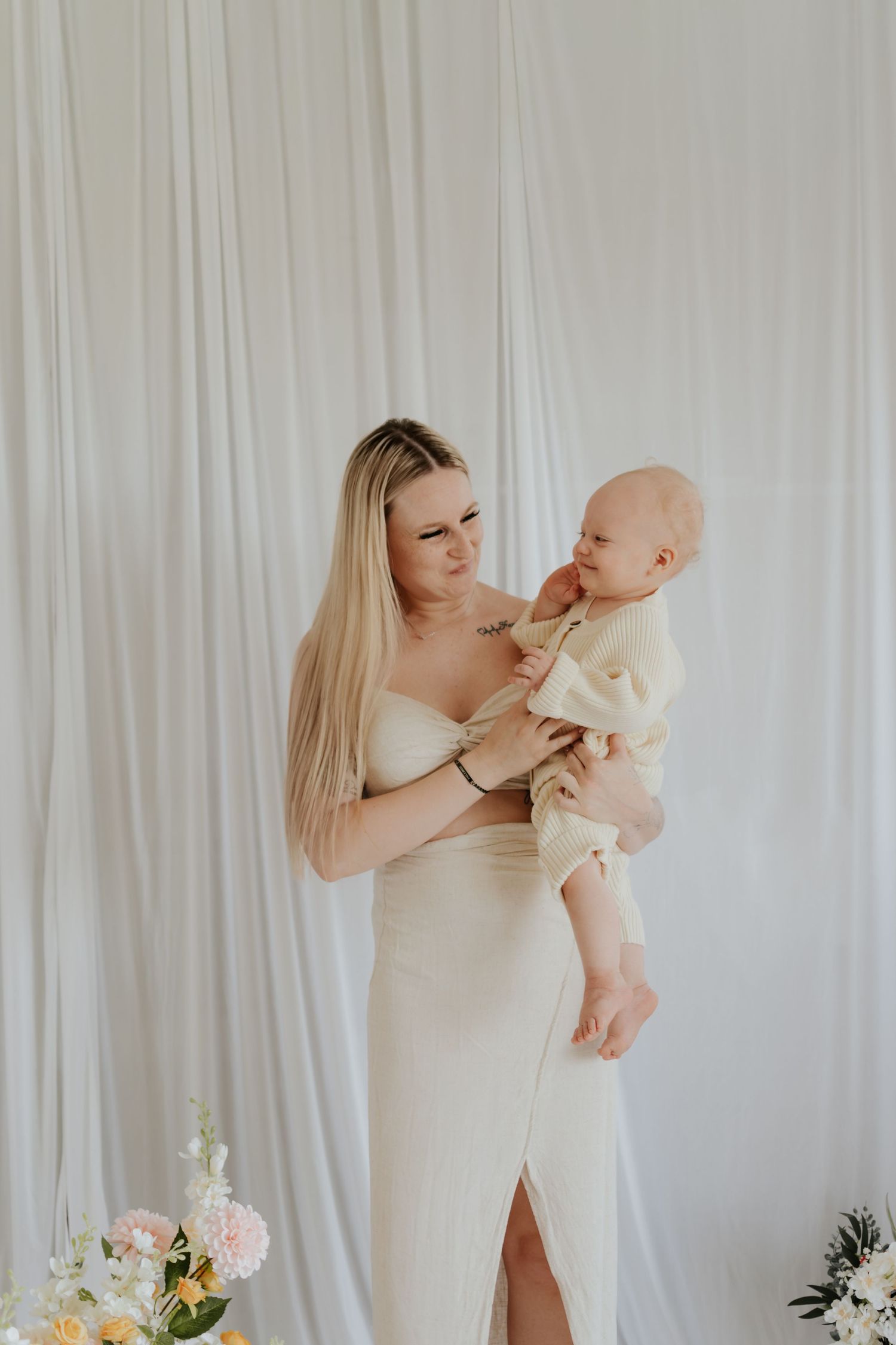 A young blonde woman in a cream dress holds a baby in a soft yellow outfit against a white draped backdrop.