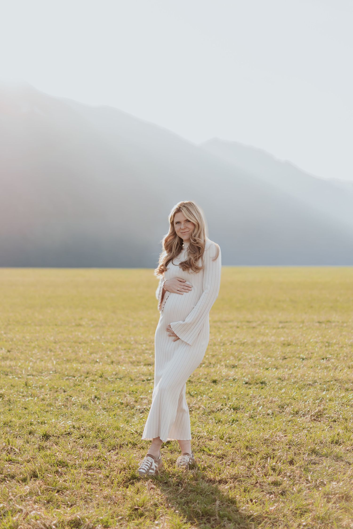 Pregnant woman in white dress standing in sunlit field with mountains in background, cradling baby bump.