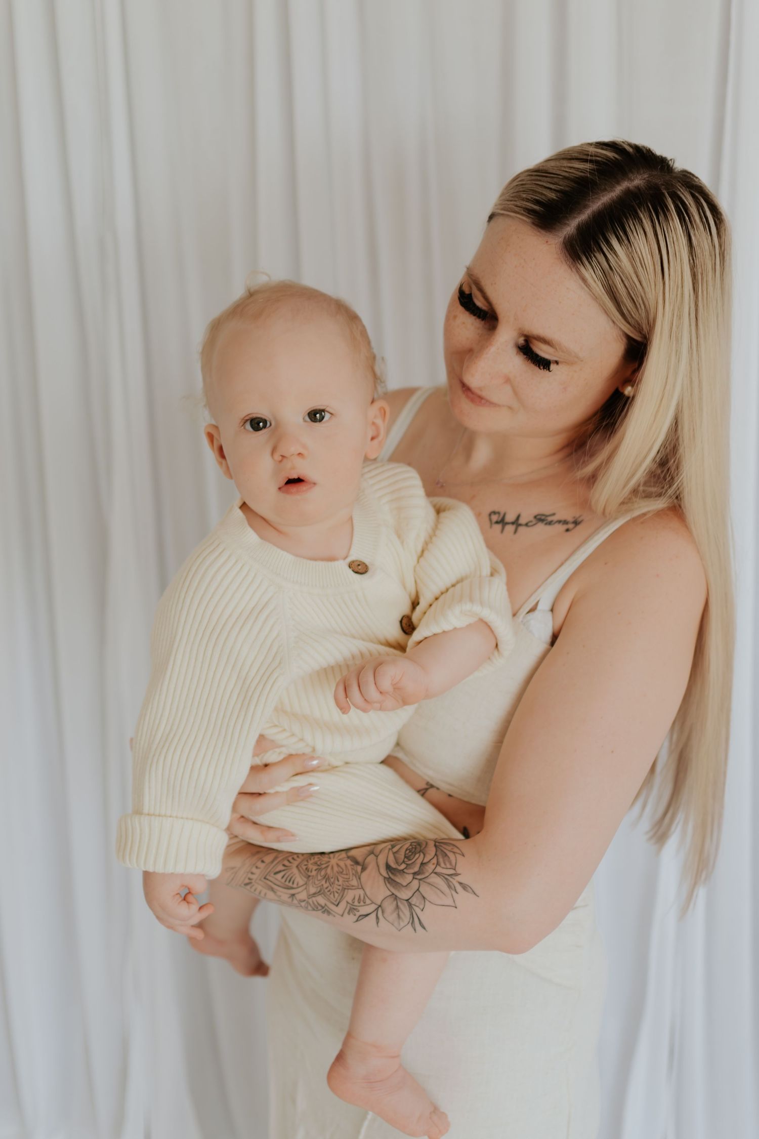 Tattooed blonde woman in cream outfit holding a baby dressed in a knit sweater against a white curtain backdrop.