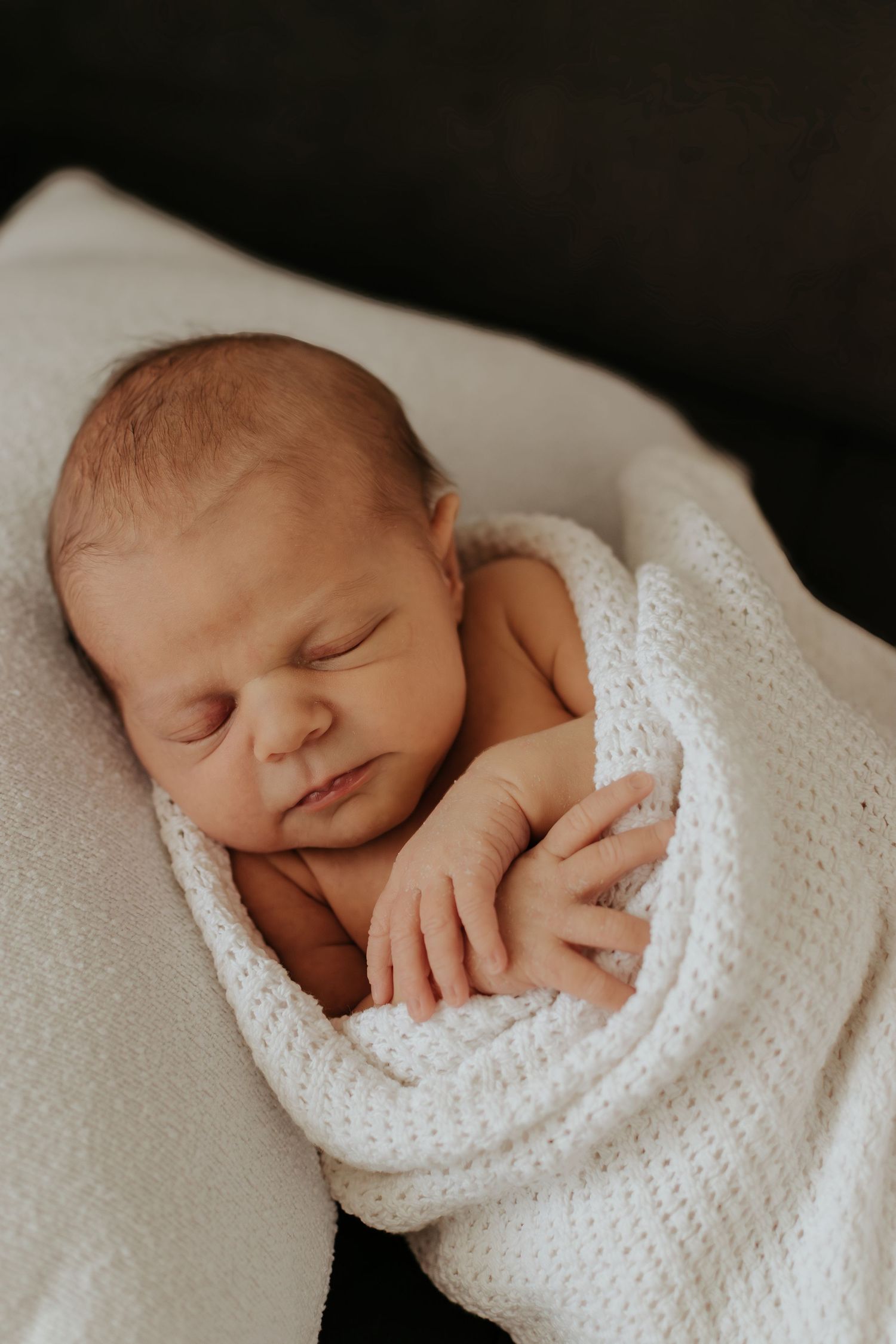 Peaceful newborn baby sleeping soundly wrapped in a soft white knit blanket with tiny hands visible.