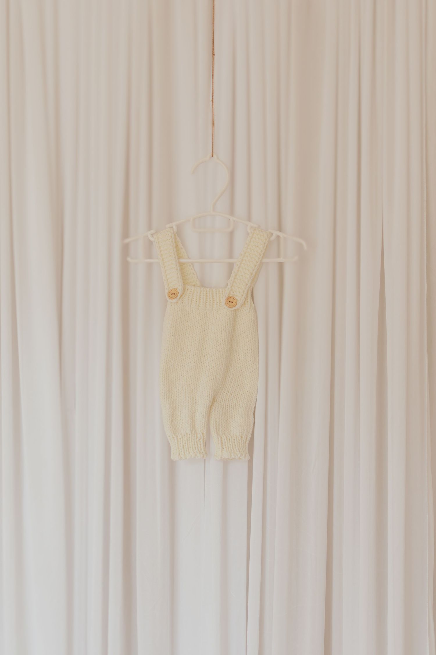 Weißes Strickoutfit für Neugeborene auf vertikalen weißen Vorhängen mit Lichtfilterung, die ein weiches, ätherisches Muster aus parallelen L