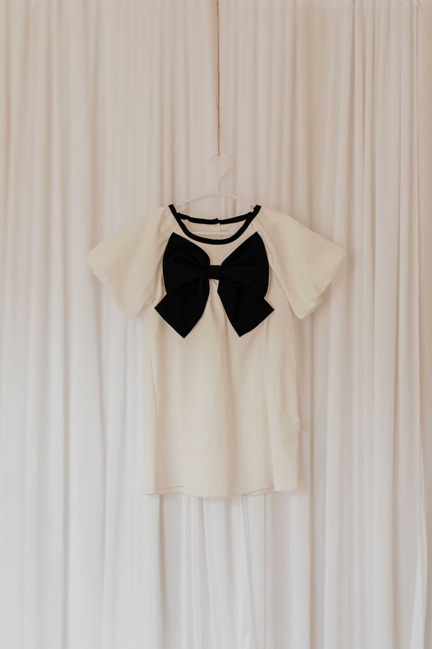Weißes Kleid mit großer schwarzer Fliege am Ausschnitt vor einem weißen Vorhanghintergrund.