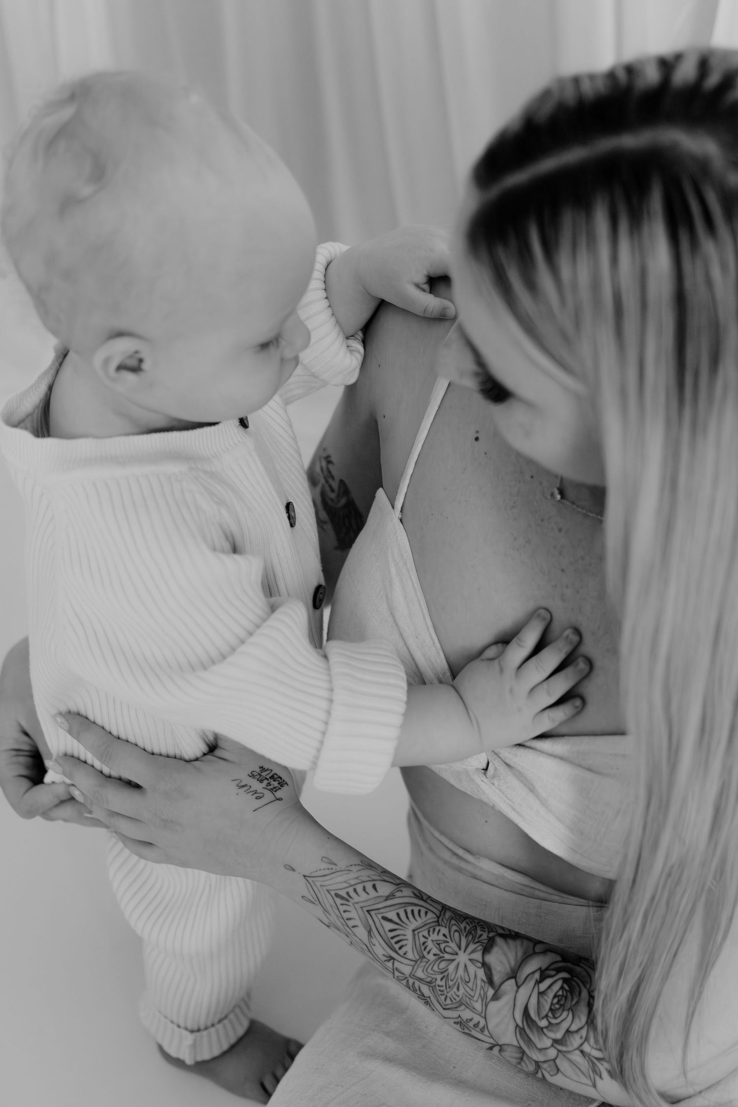 Black and white photo of a mother holding her baby, both sharing a tender moment, with tattoos visible on her arm.