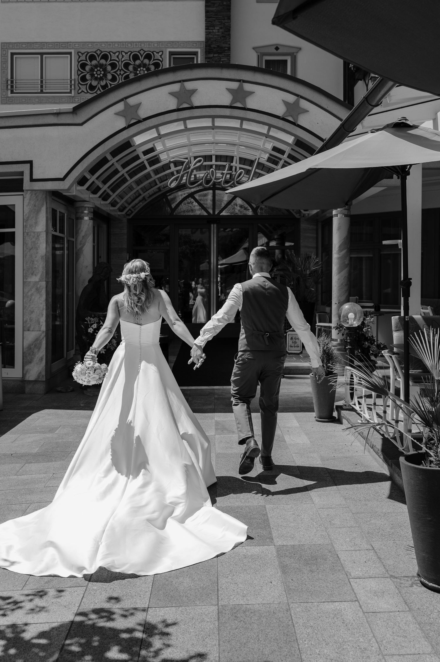 Black and white photo of a bride and groom holding hands walking toward a hotel entrance on their wedding day.