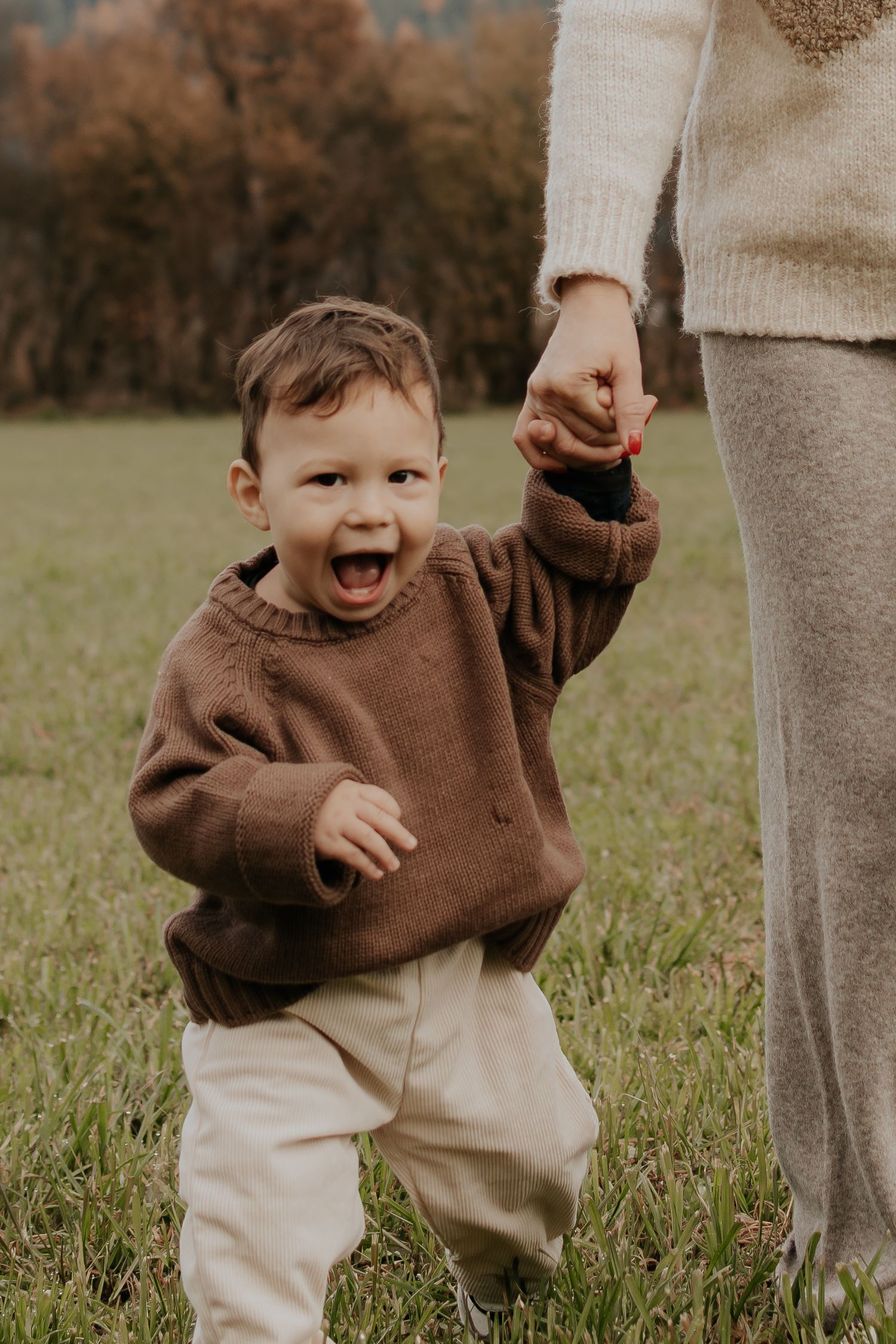 Energiegeladenes Kleinkind im braunen Pullover spielt mit einem Familienmitglied in einem cremefarbenen Outfit.