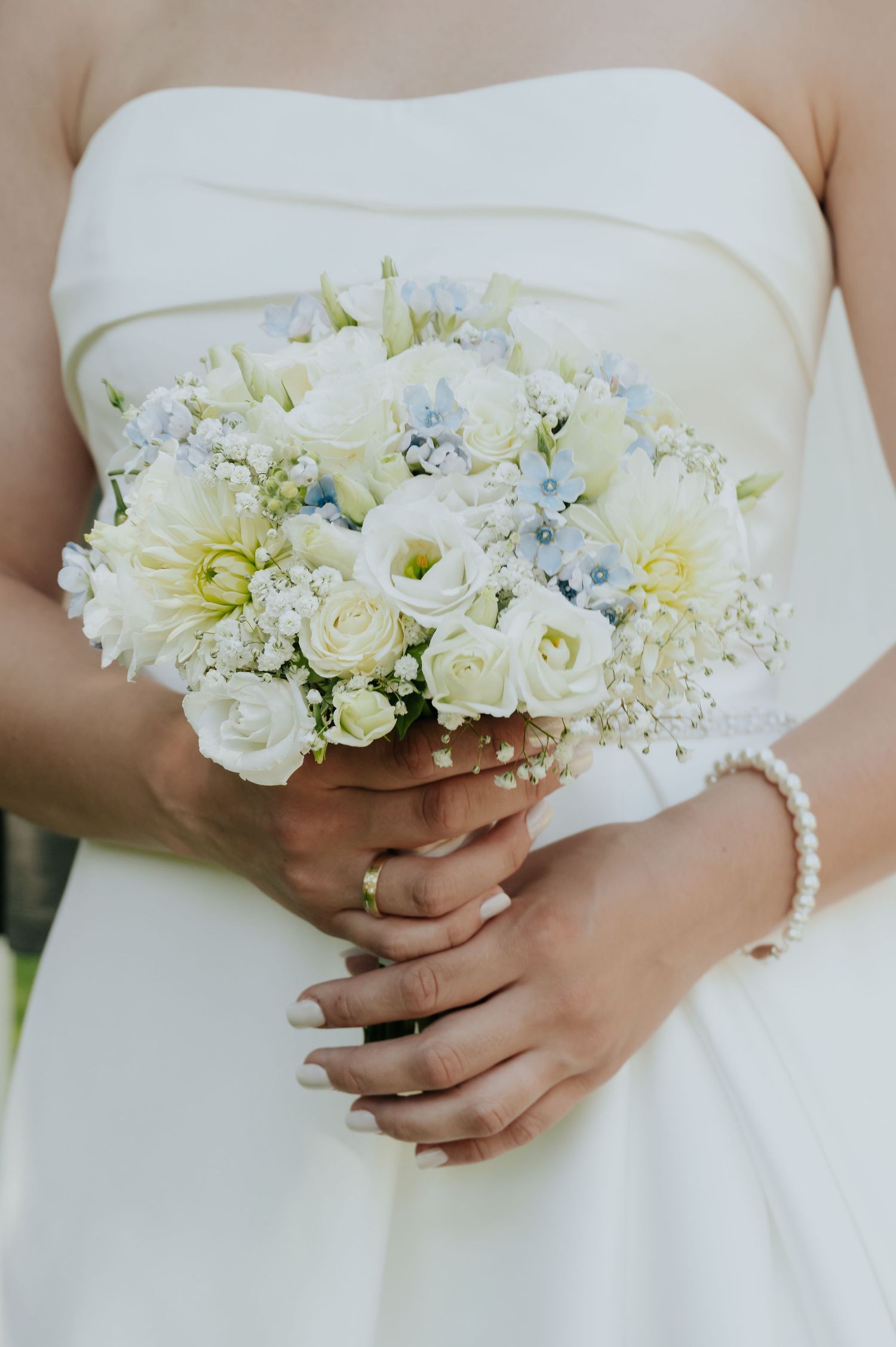 Bride in strapless white gown holds elegant bouquet of white roses, dahlias, and blue flowers with pearl bracelet.
