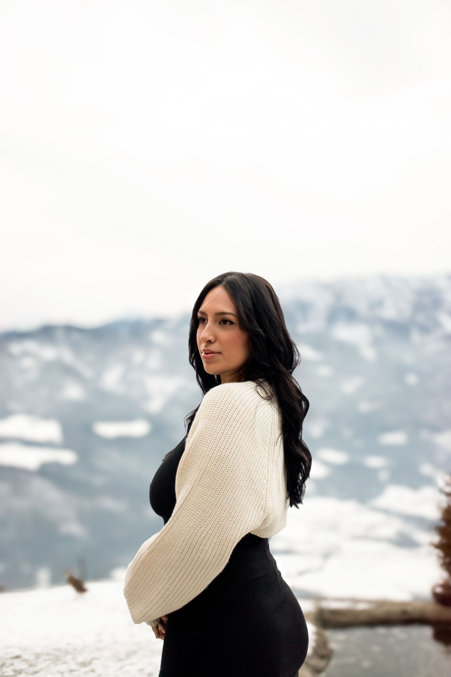 Frau im cremefarbenen Pullover posiert vor verschneiter Berglandschaft.