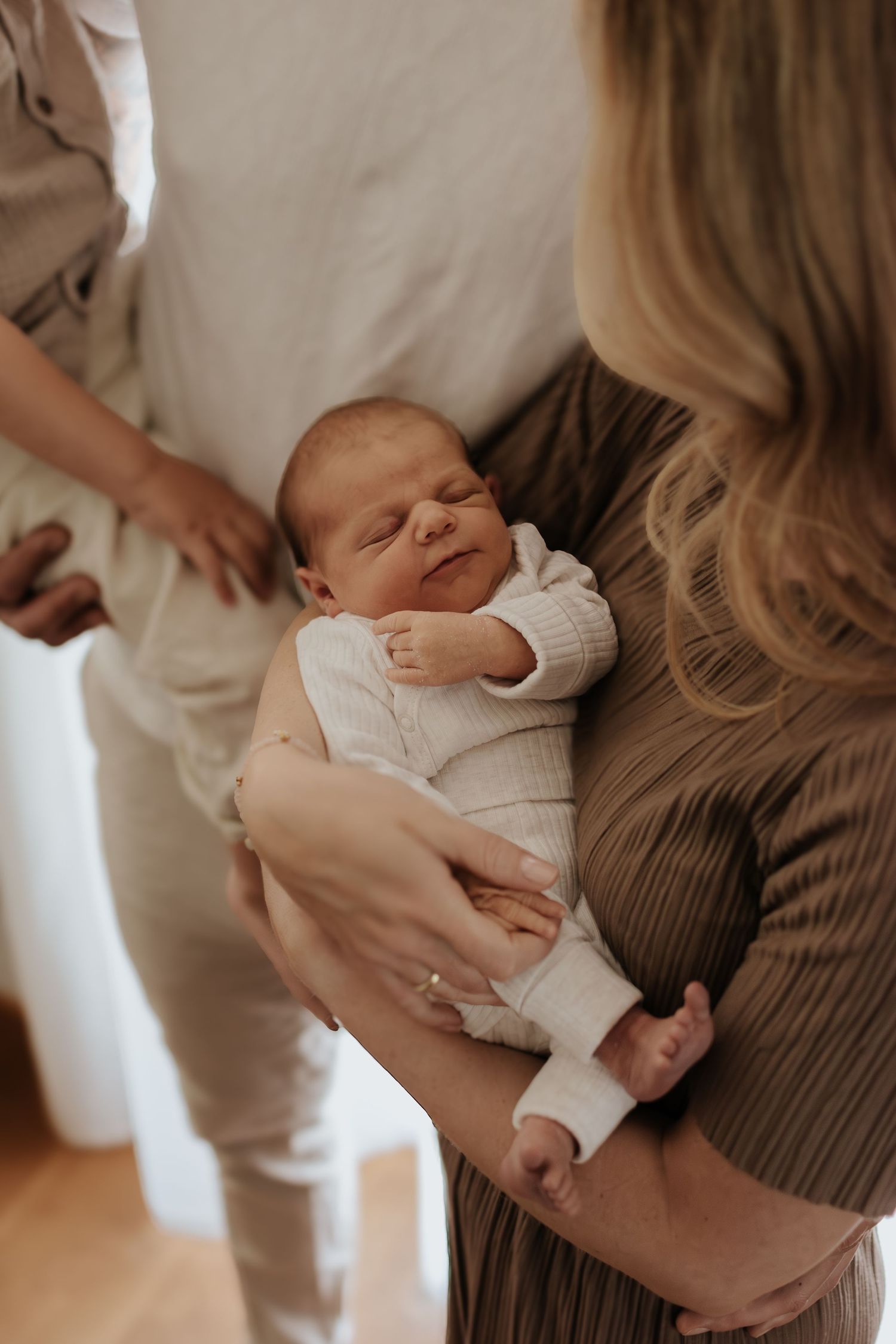 A newborn baby sleeping peacefully in a woman's arms, dressed in a white knit outfit, surrounded by family.