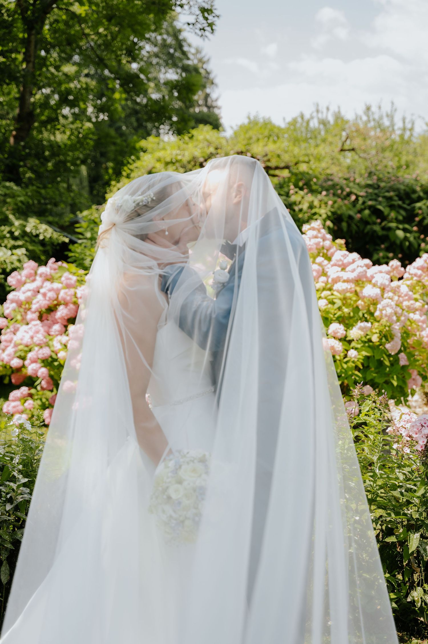 Bride and groom sharing a romantic kiss hidden beneath a flowing white veil in a flower garden.