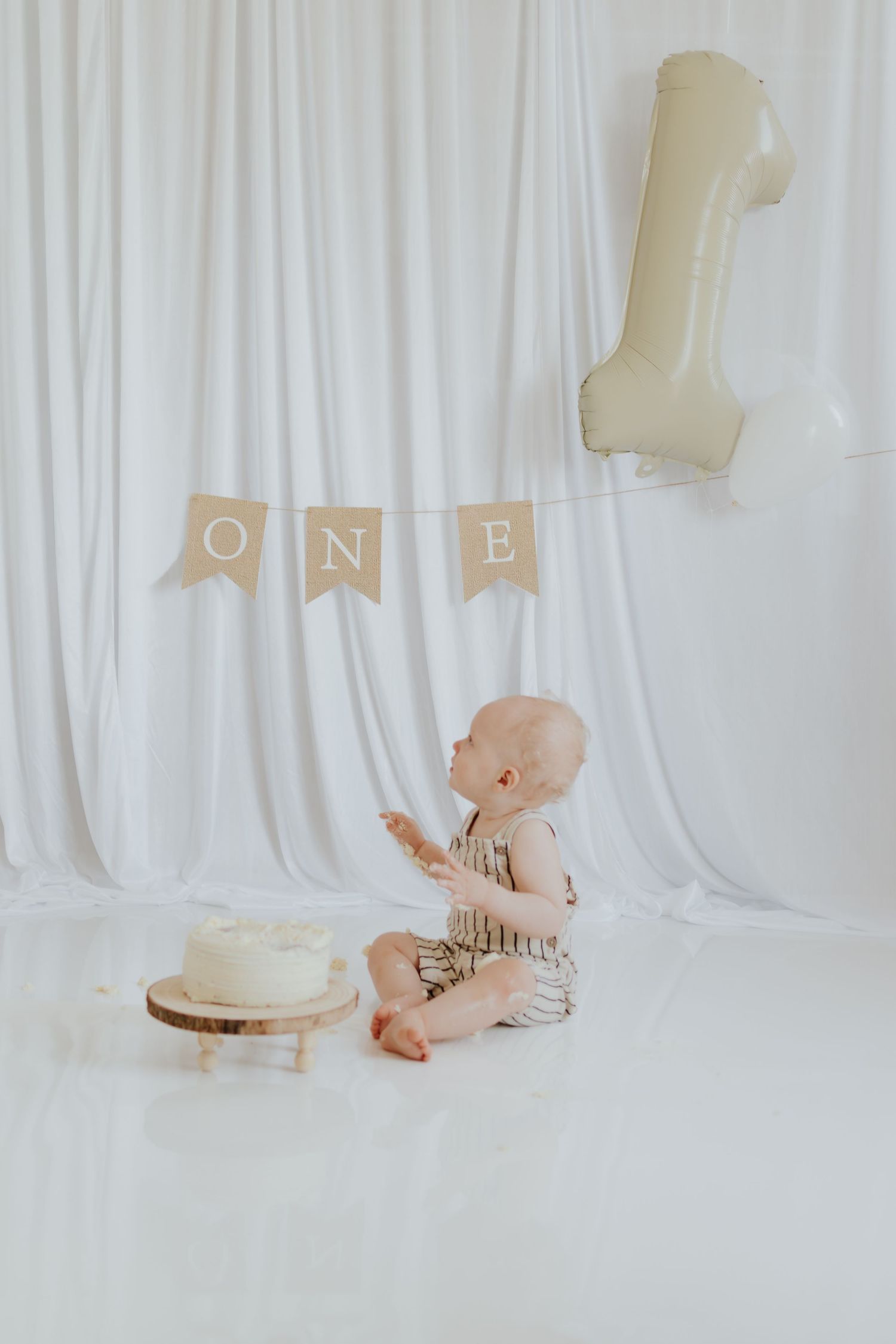Baby's first birthday cake smash with white cake, ONE banner, and number 1 balloon on white backdrop.