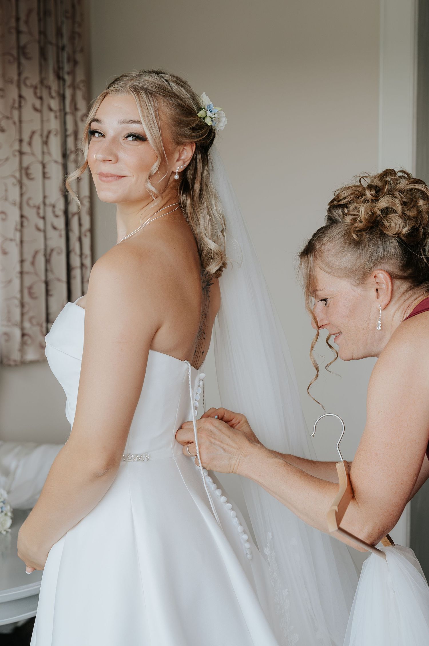 Bride in strapless white gown smiles over shoulder as woman fastens buttons on wedding dress back.