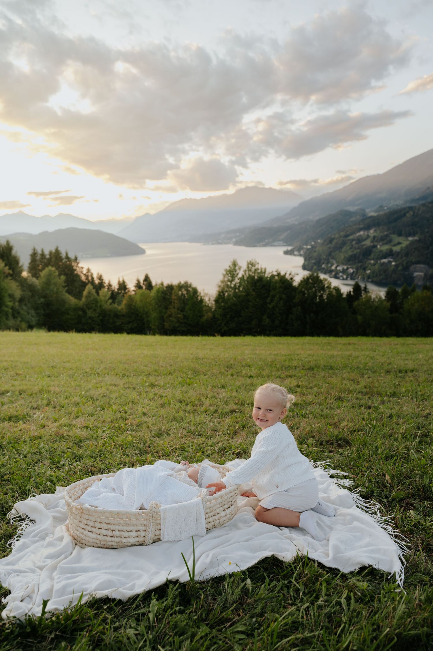 Baby in white outfit sits on blanket with wicker basket, smiling before scenic mountain lake at sunset.