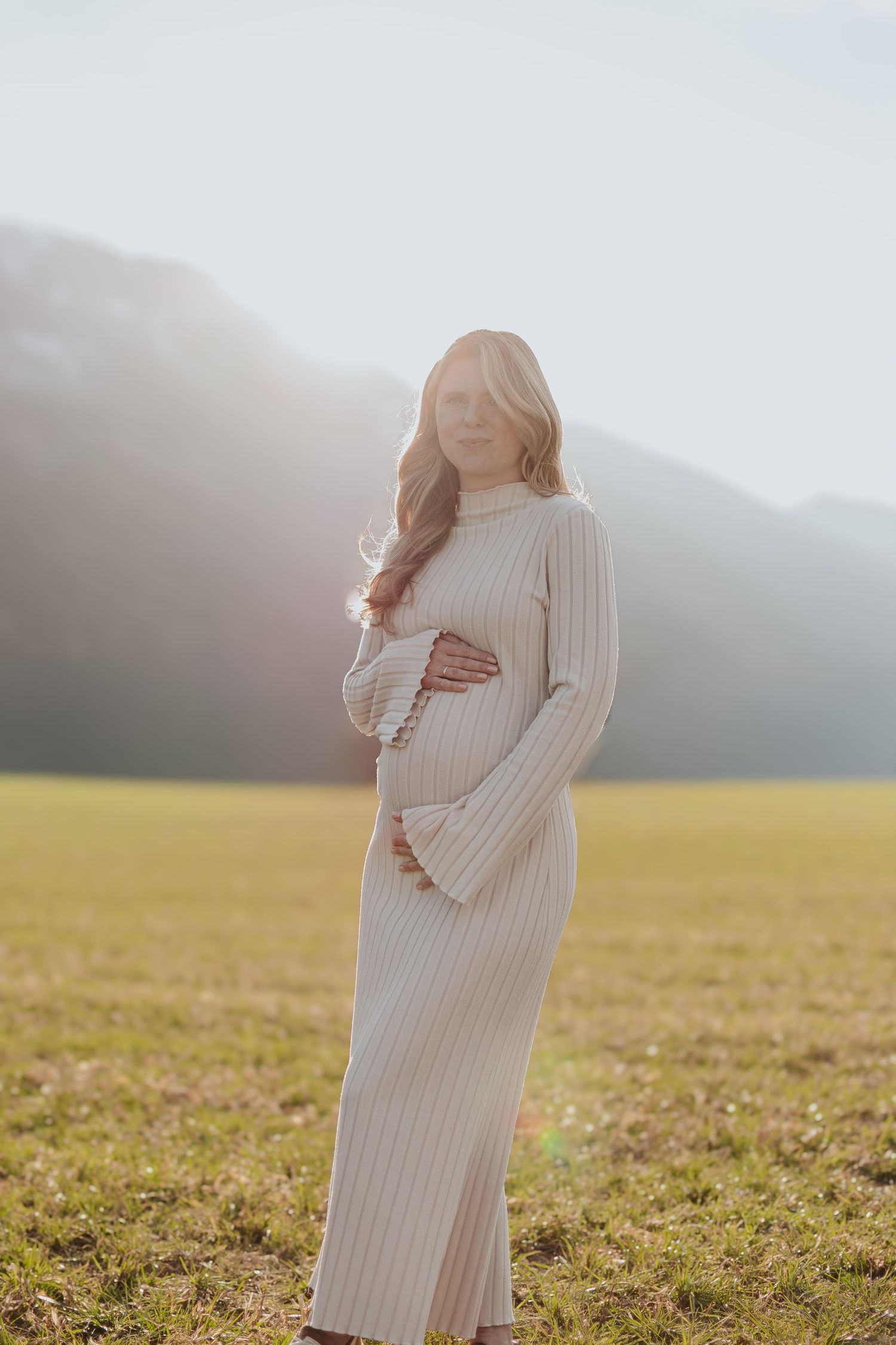 Pregnant woman in cream ribbed dress standing in sunlit field with misty mountains behind.