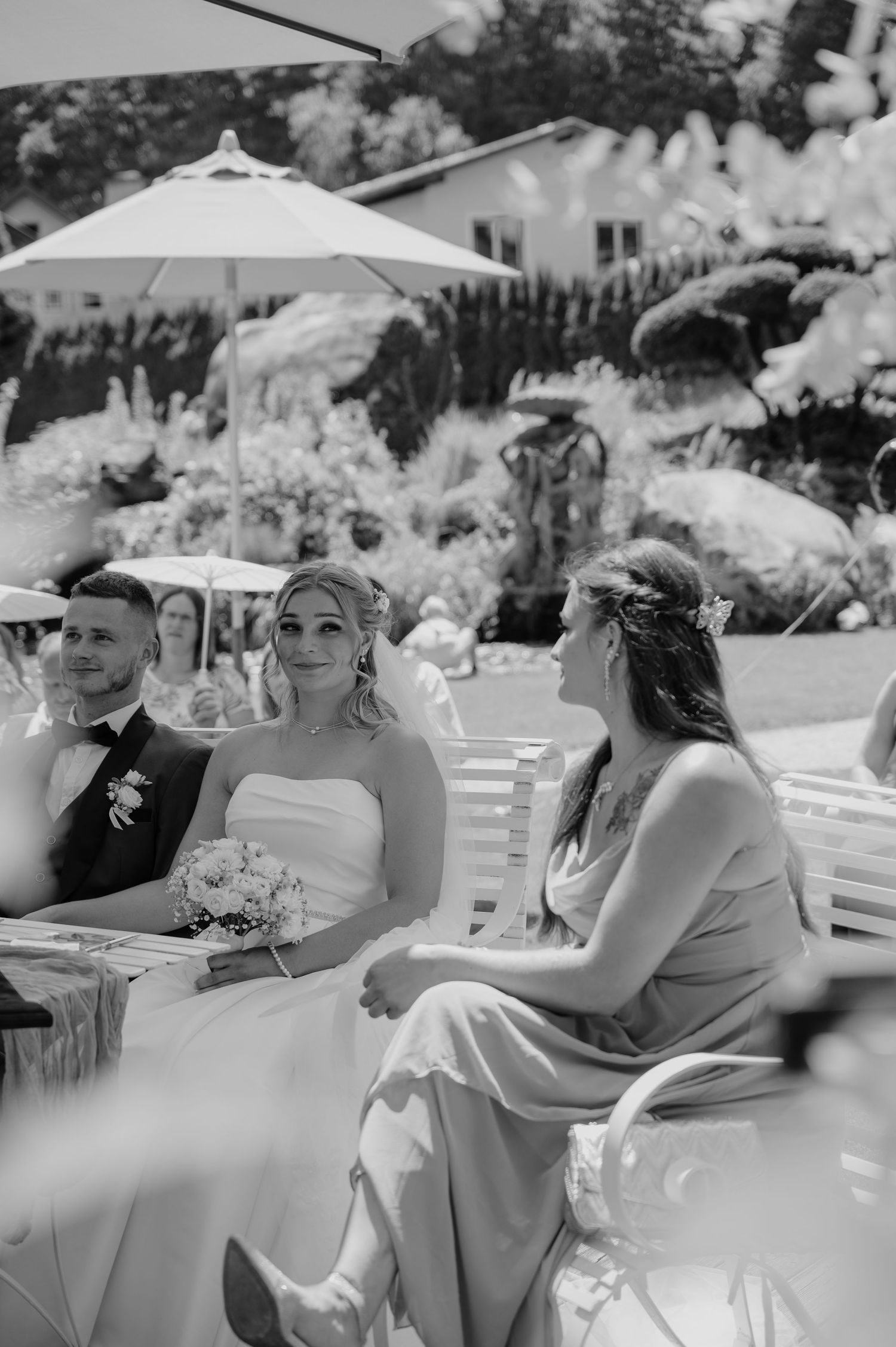 Black and white photo of a bride in white dress holding bouquet, seated with groom and bridesmaid at outdoor wedding ceremony.