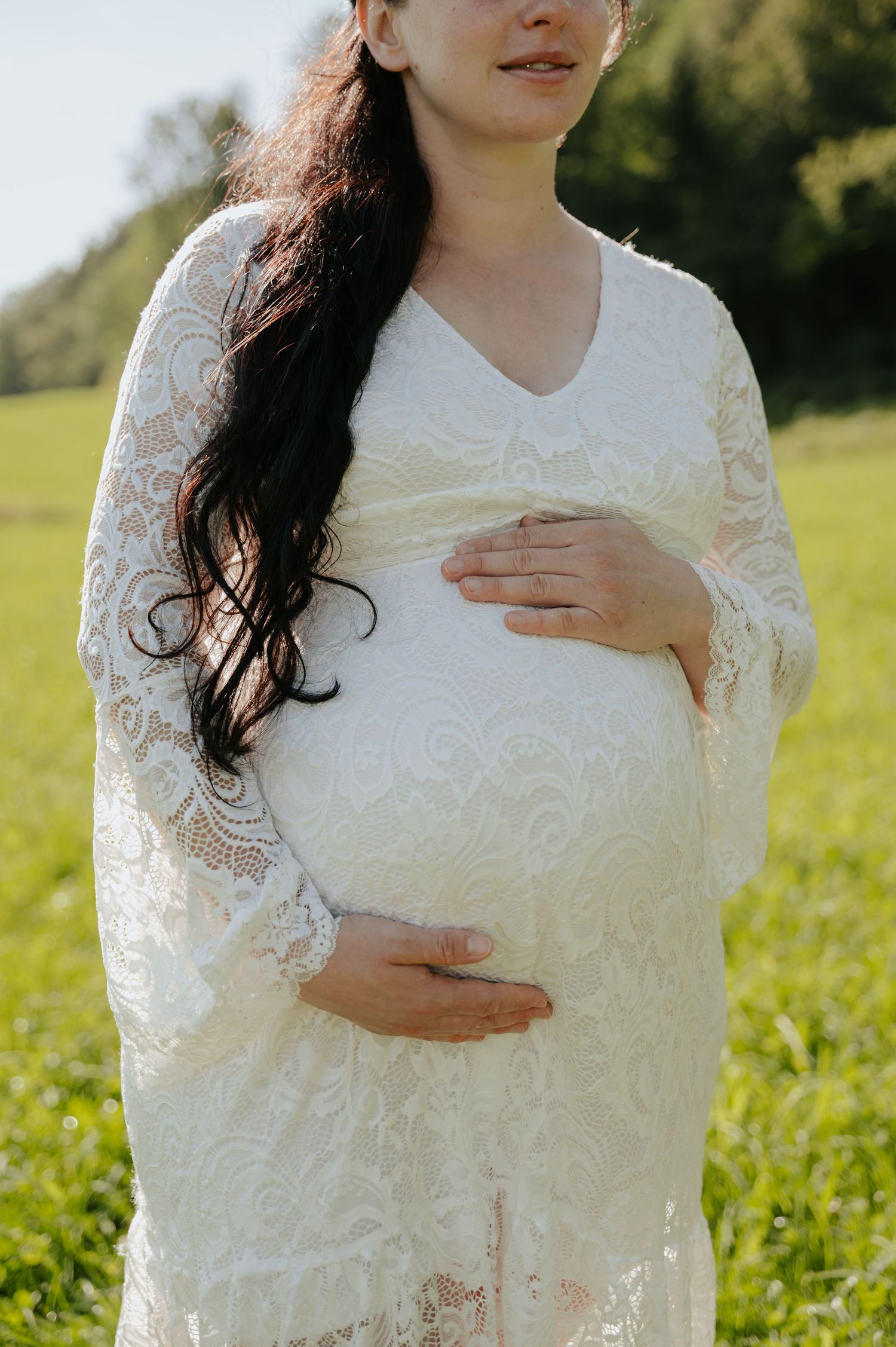 Pregnant woman in white lace dress cradling baby bump outdoors in green field during golden hour.