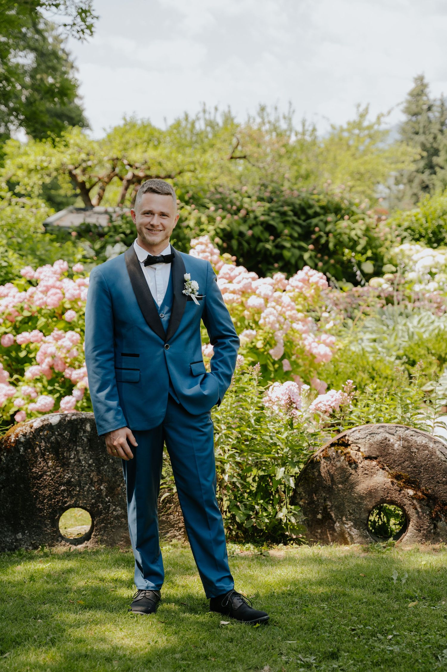 Handsome groom in blue tuxedo with bow tie posing in a lush garden with pink hydrangeas.