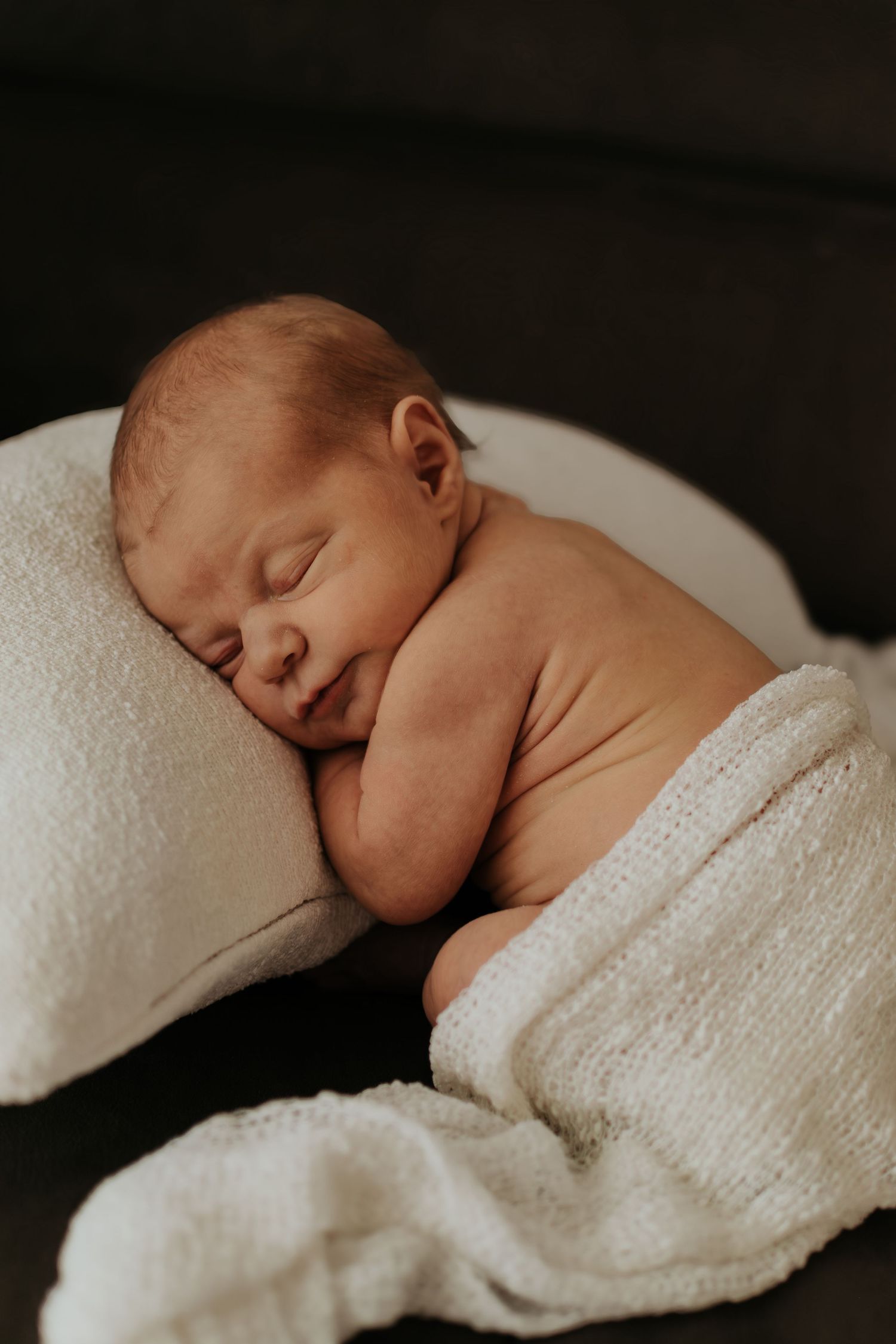 A peaceful newborn baby sleeping on a white pillow wrapped in a soft white blanket against a dark background.