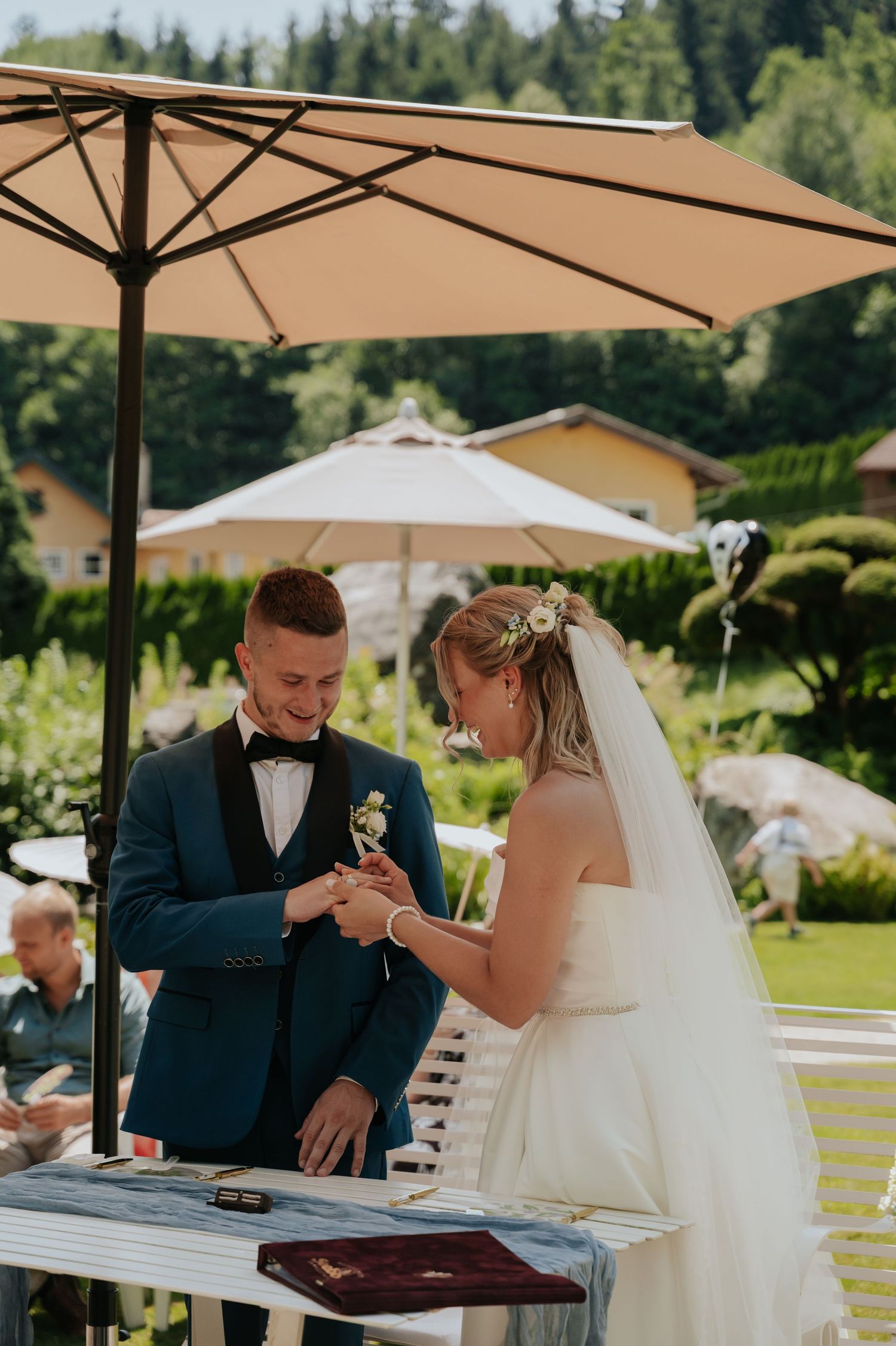 Bride placing wedding ring on groom's finger outdoors under a patio umbrella during a sunny garden ceremony.