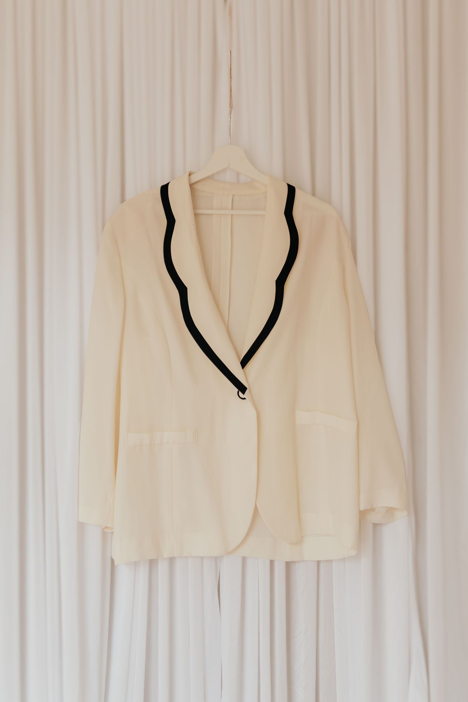 Ein cremefarbener Blazer mit schwarzem Rand hängt vor einem weißen Vorhanghintergrund.