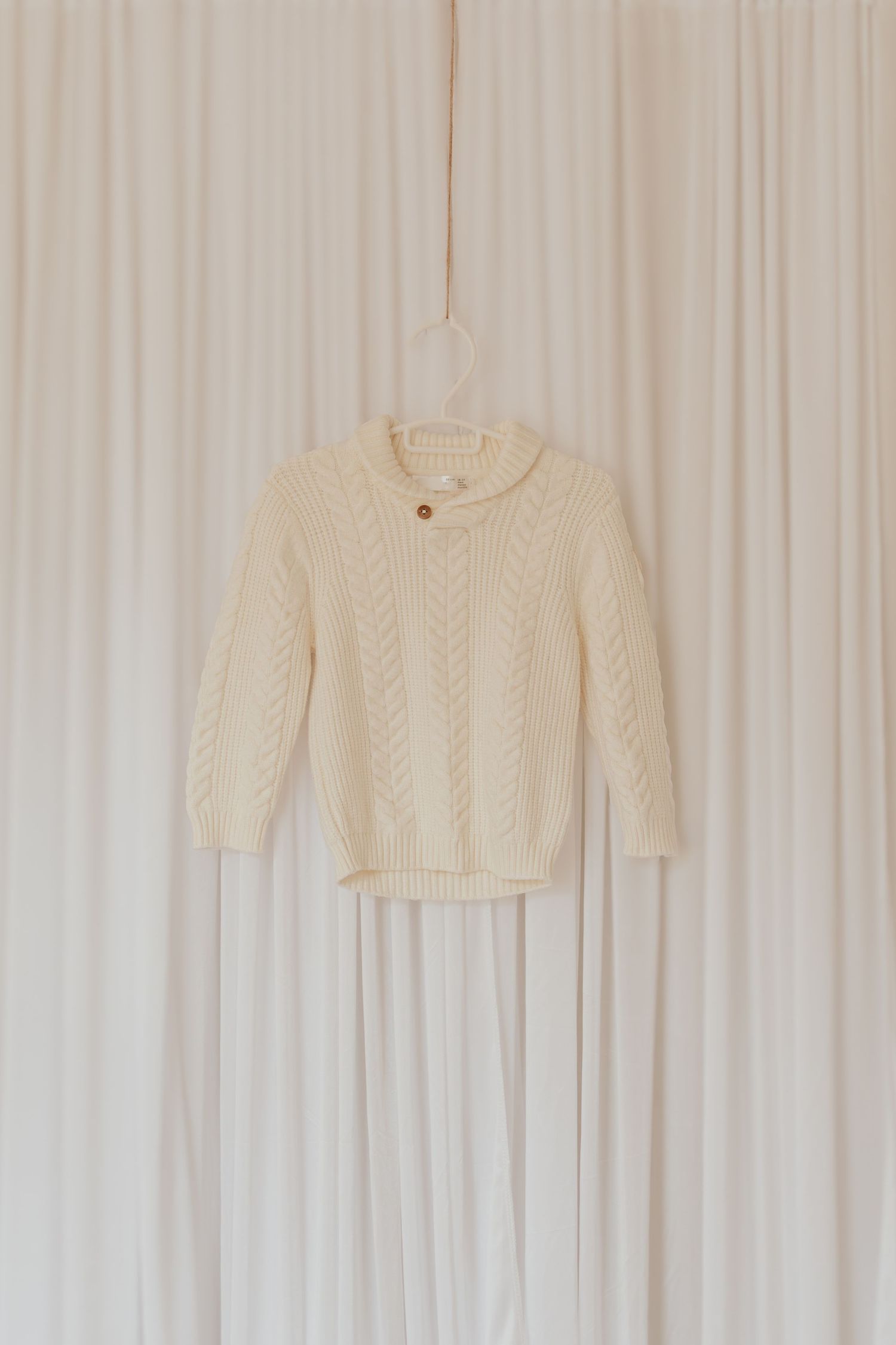 Cremefarbener Kabelstrickpullover, der auf weißem Vorhanghintergrund mit weichem natürlichem Licht hängt.