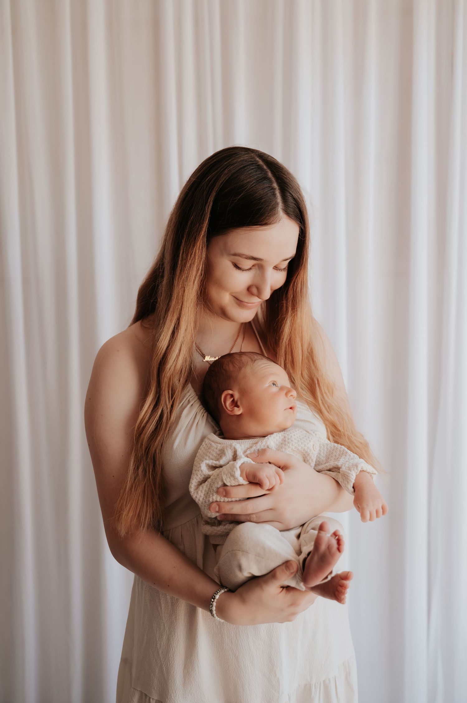 A young mother in a white dress tenderly holds her sleeping newborn baby against a soft white curtain backdrop.
