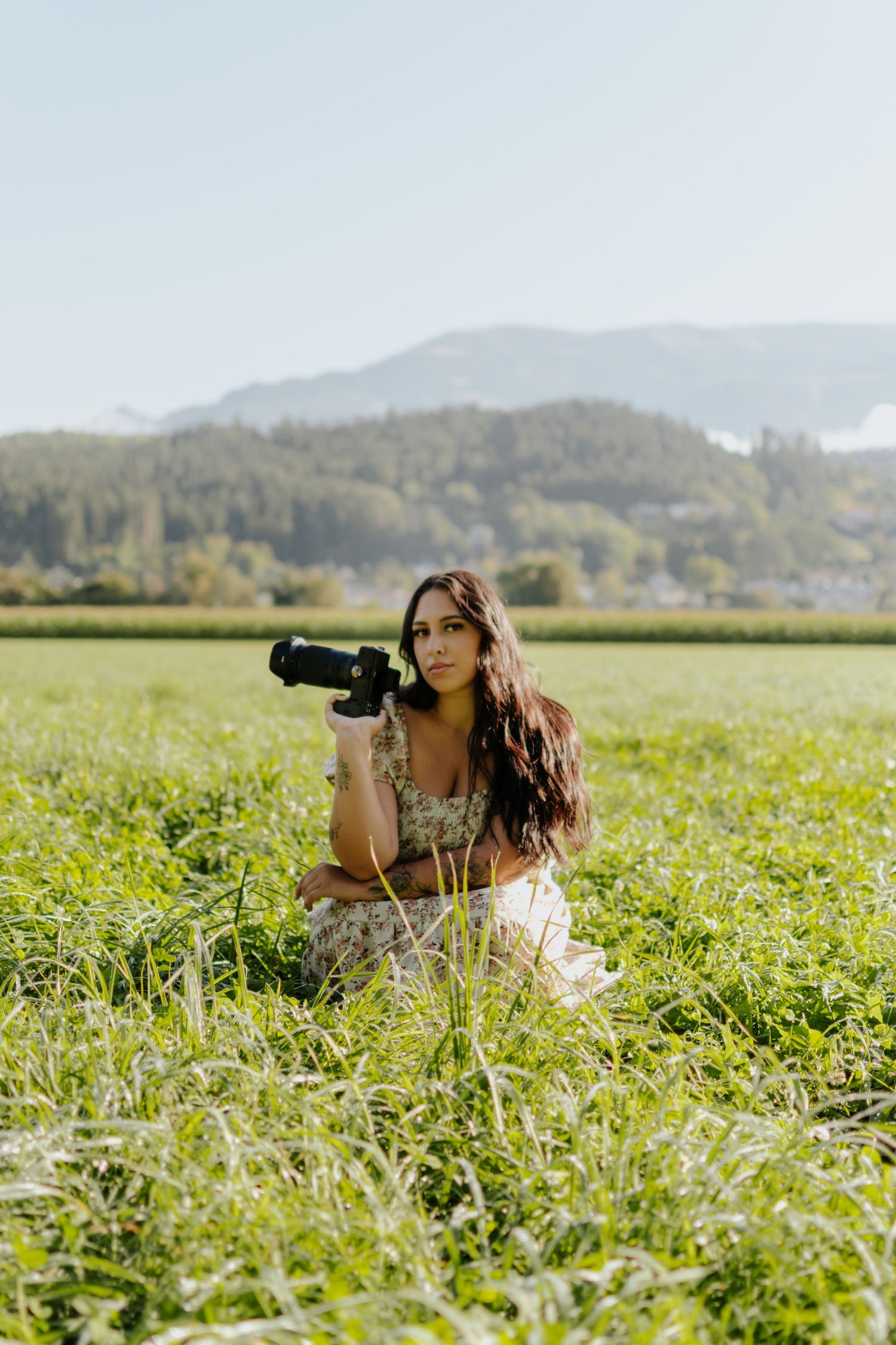Woman in floral dress kneeling in green field holding camera with mountain backdrop on sunny day.