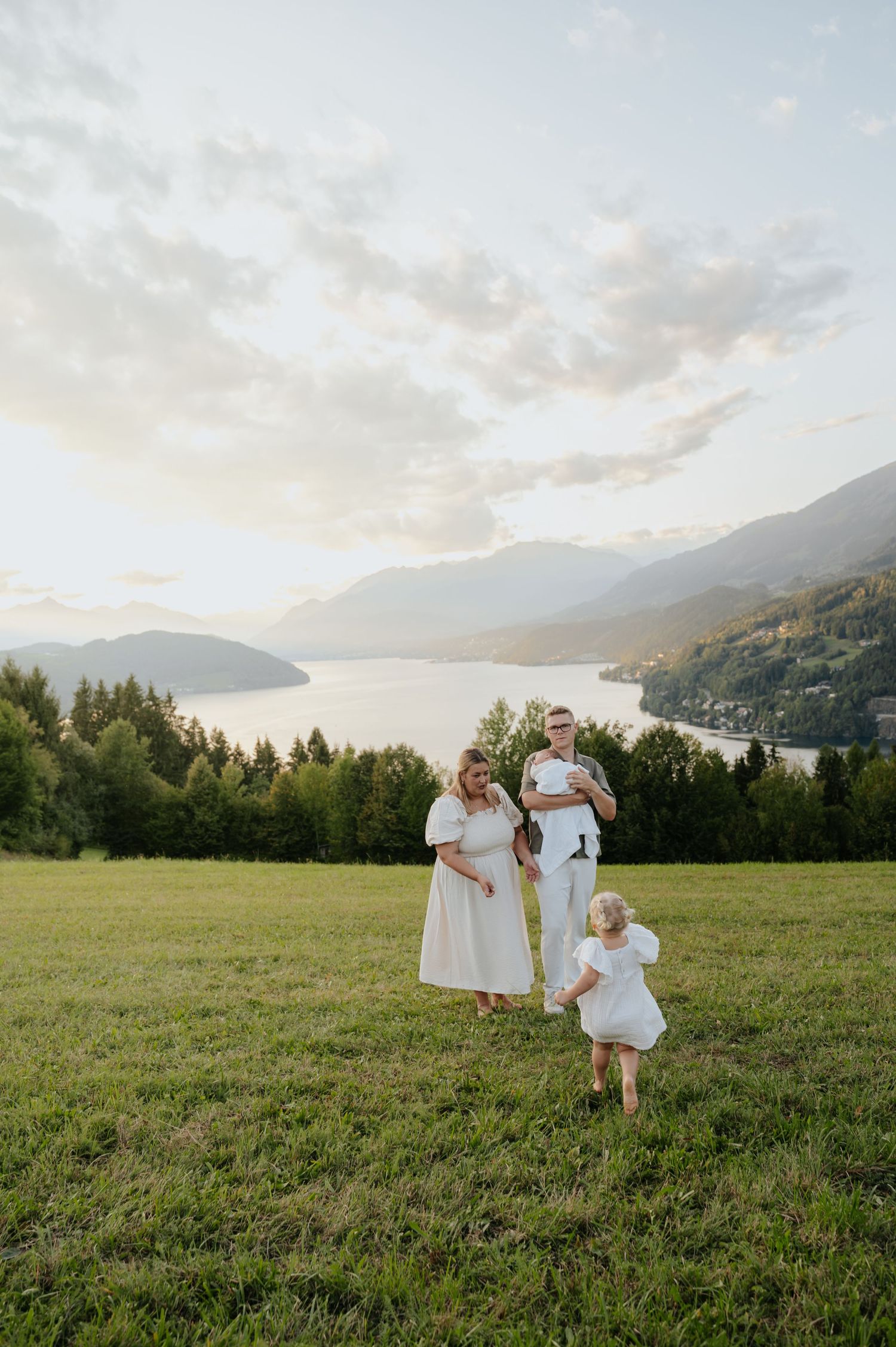 Family in white outfits posing on green hillside with mountain lake and forest backdrop at golden hour.