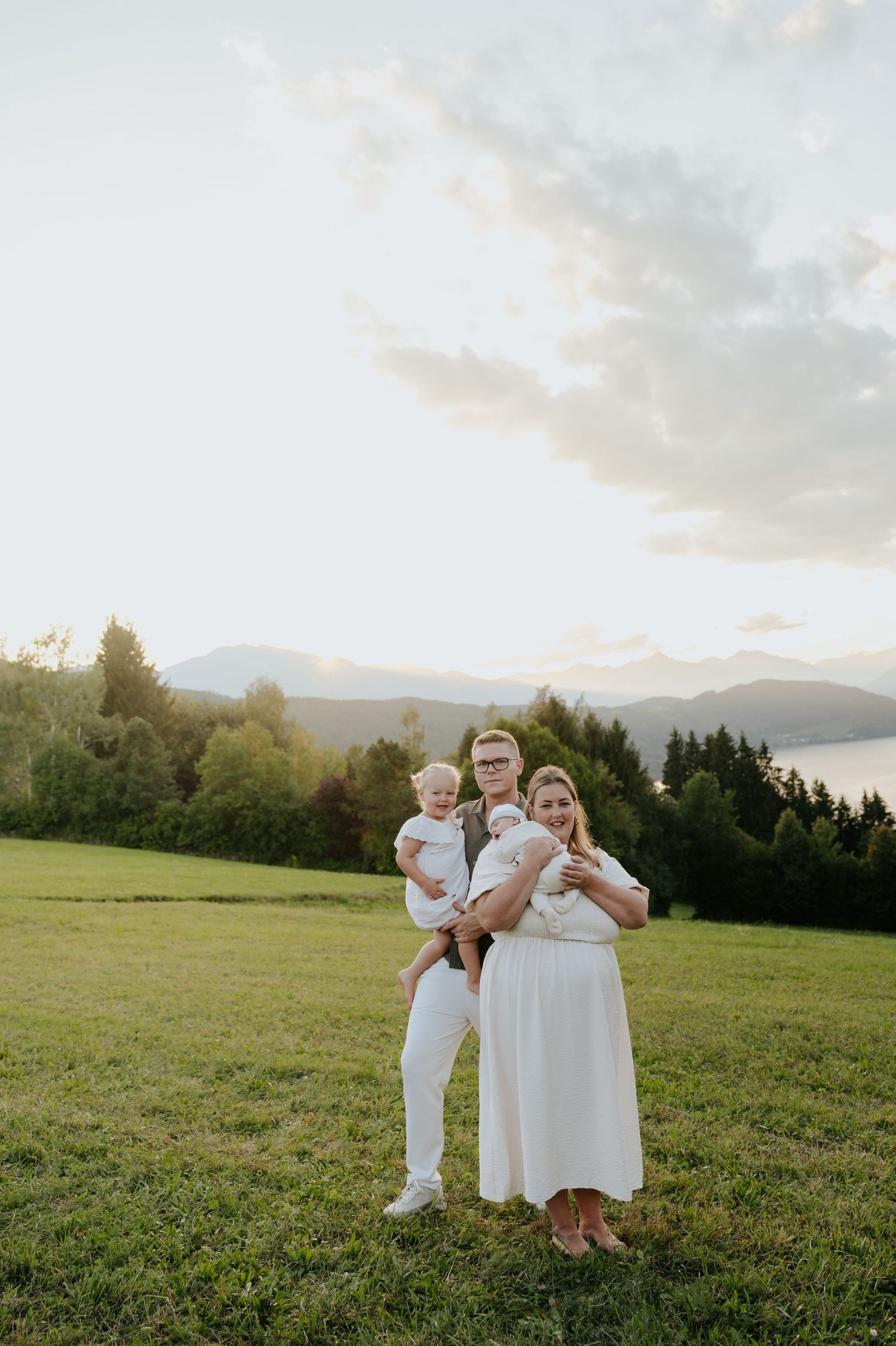 Family of three in white outfits posing on green meadow with mountain lake and forest backdrop at golden hour.