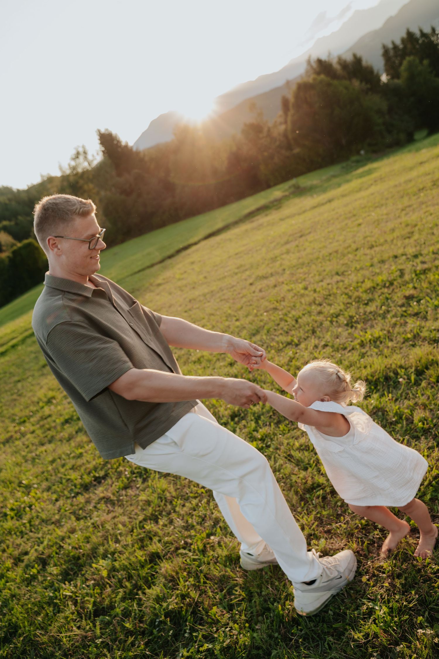 Father playfully spinning toddler daughter on sunny green field with mountain backdrop at sunset.
