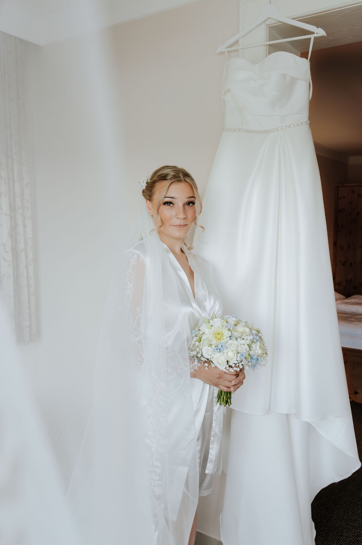 Bride in white robe holding bouquet of white and blue flowers stands beside her wedding dress hanging on a hanger.