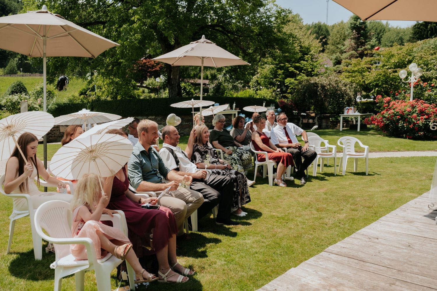 Guests seated outdoors at a sunny garden wedding ceremony, holding parasols and enjoying the celebration.